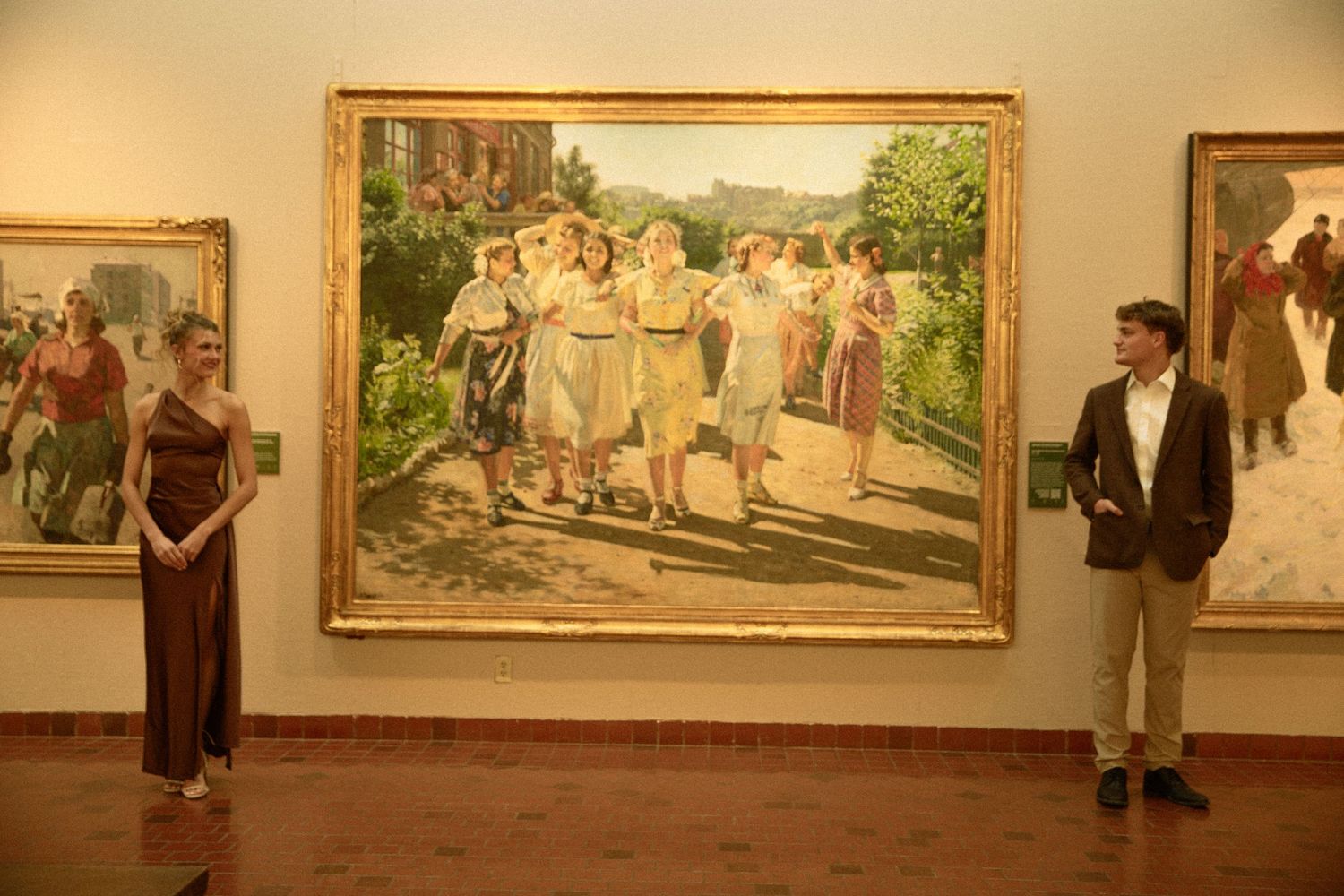 Museum visitors observe a large painting depicting people in white dresses walking along a path in a summer garden scene.