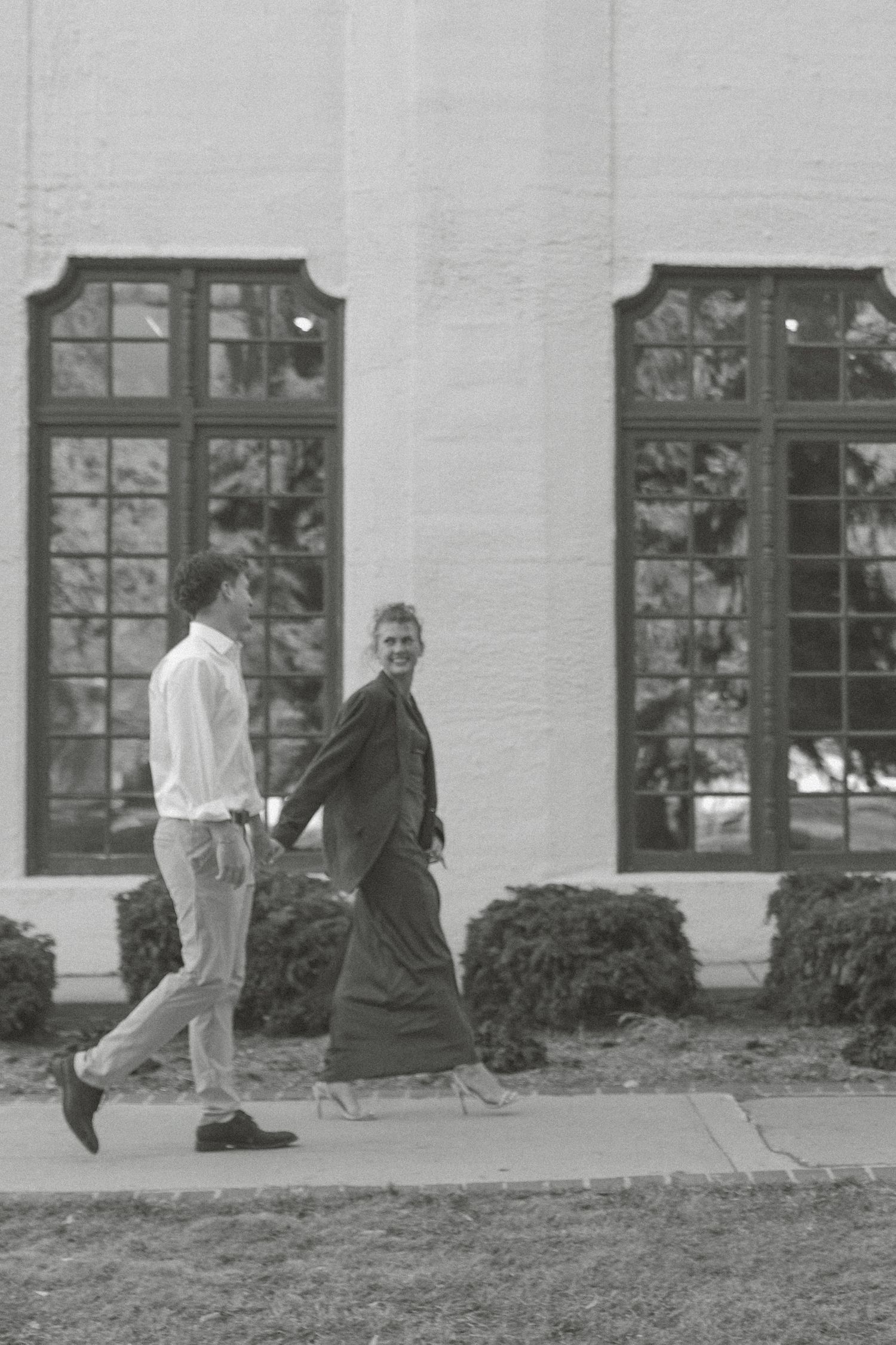 A black and white photograph shows two people walking past tall windows on a building exterior.