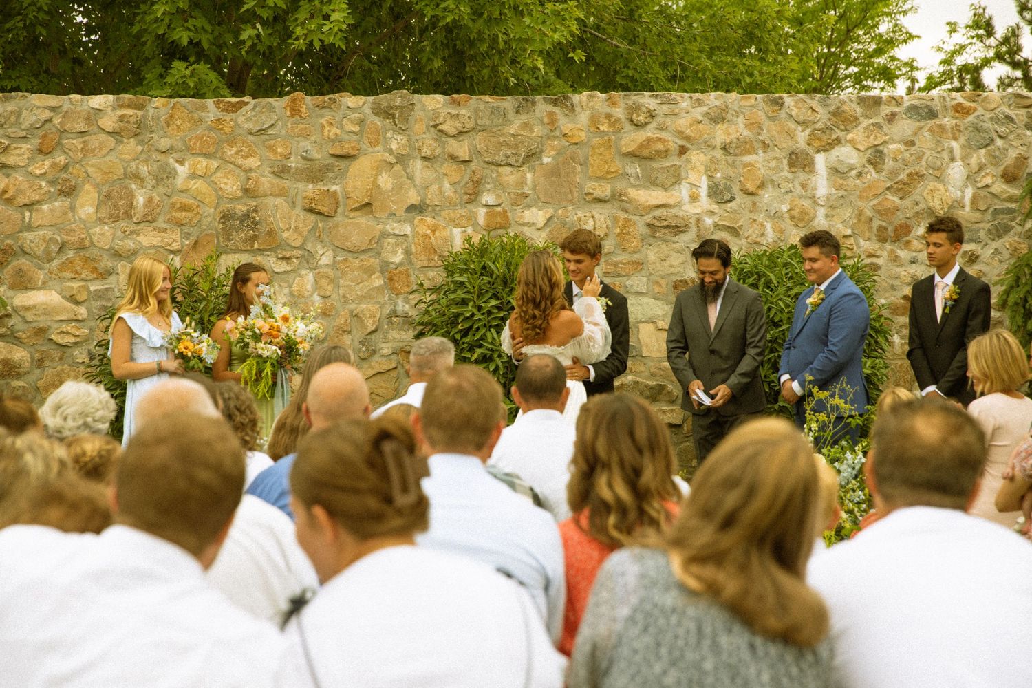 Wedding guests gather outdoors in front of a stone wall during a summer celebration.