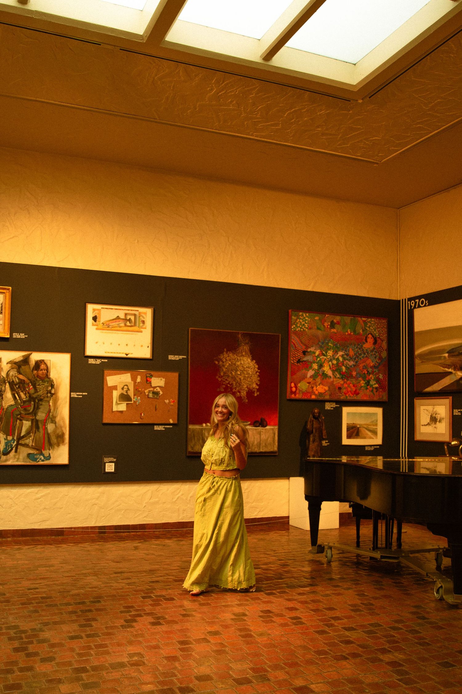 Woman in yellow dress standing in art gallery with colorful paintings on dark walls and skylight ceiling.
