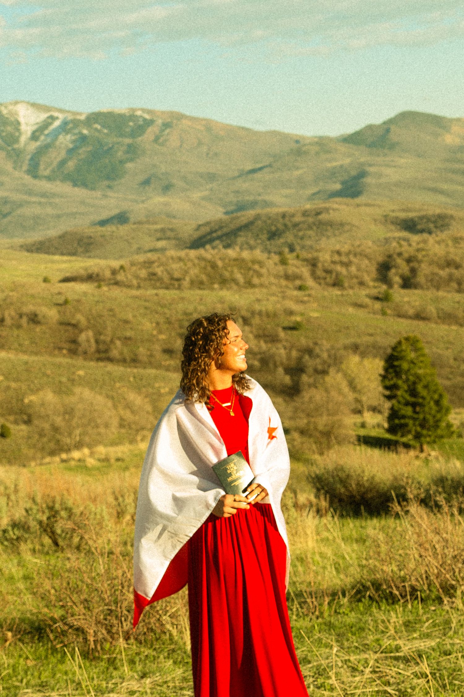 Woman in red dress and white shawl standing in green mountain meadow landscape on sunny day.