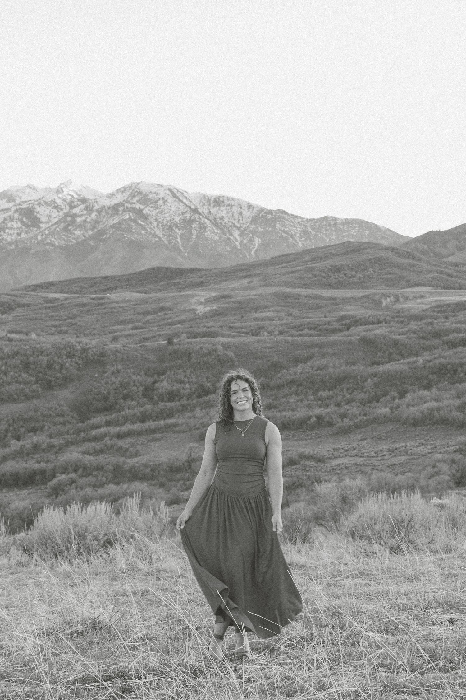 Black and white photo of a woman in a flowing dress standing in a mountain meadow landscape.