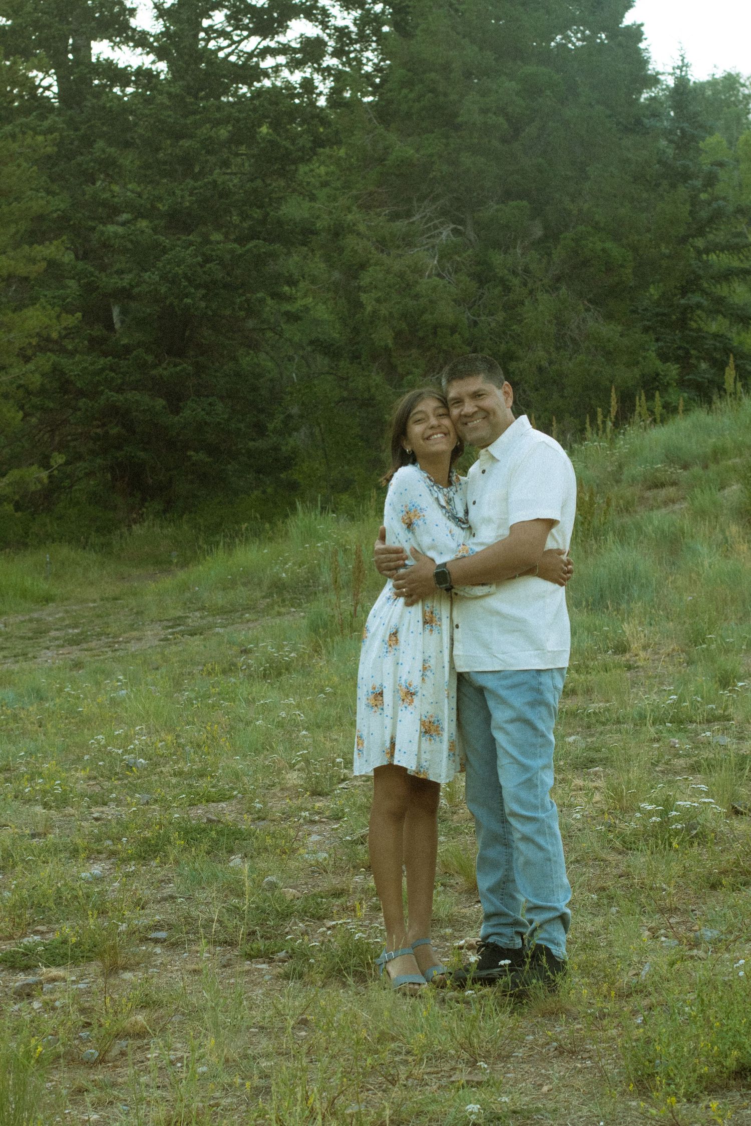 A couple embraces while standing on a grassy hillside with evergreen trees in the background.