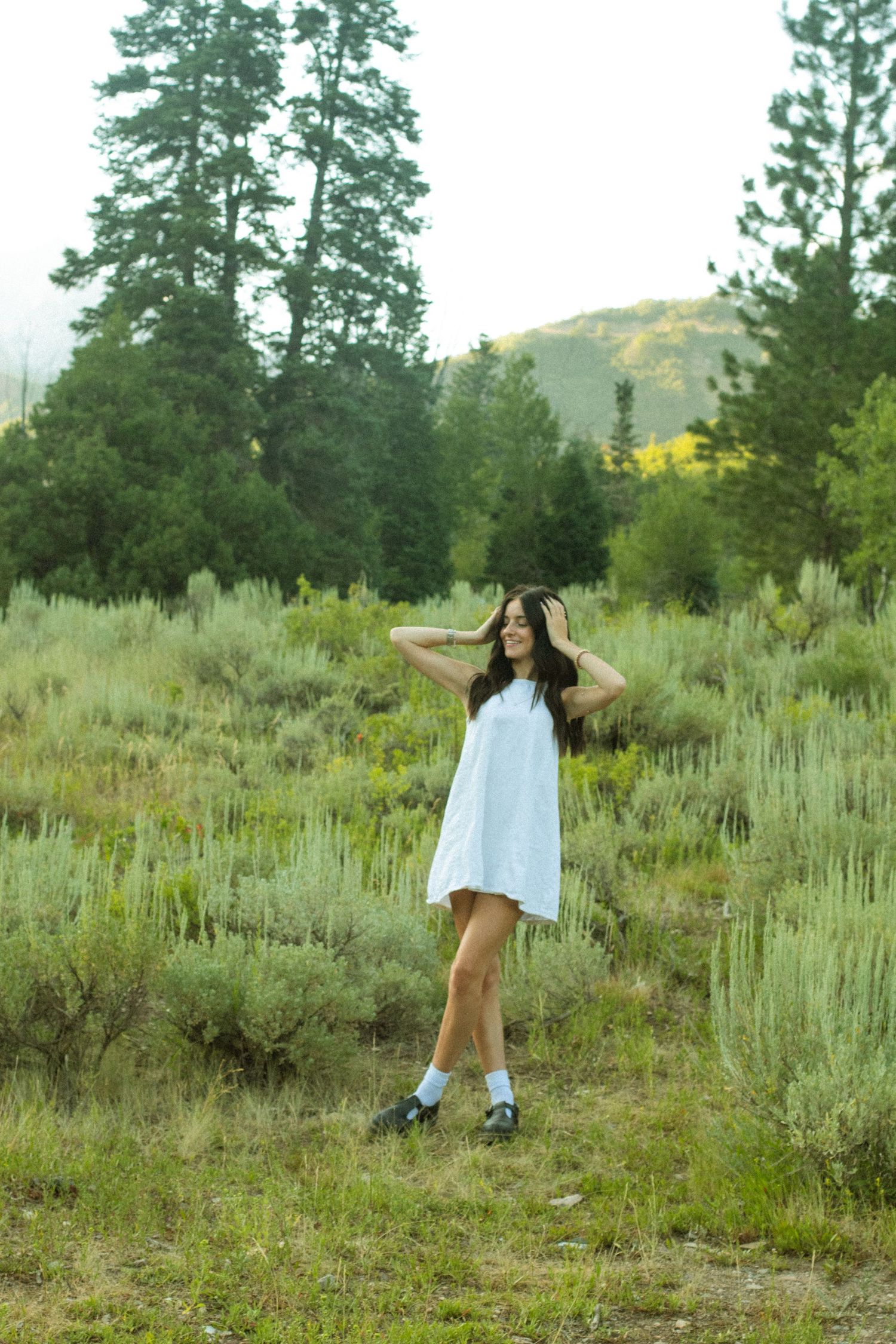 A figure in a white dress spins gracefully through a field of sagebrush and pine trees in a natural outdoor setting.