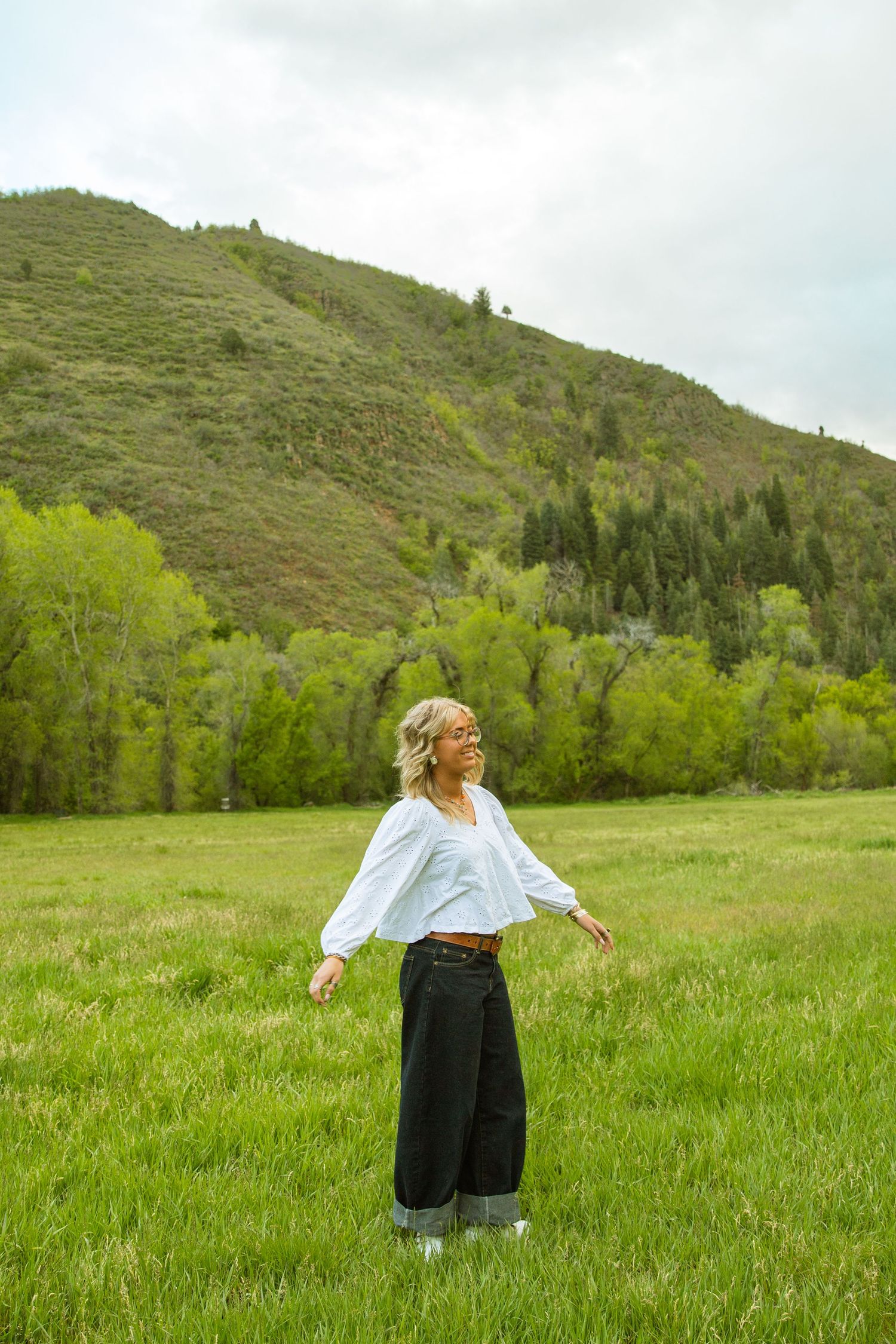 A woman in a white blouse and black wide-leg pants stands in a lush green meadow with forested hills behind her.