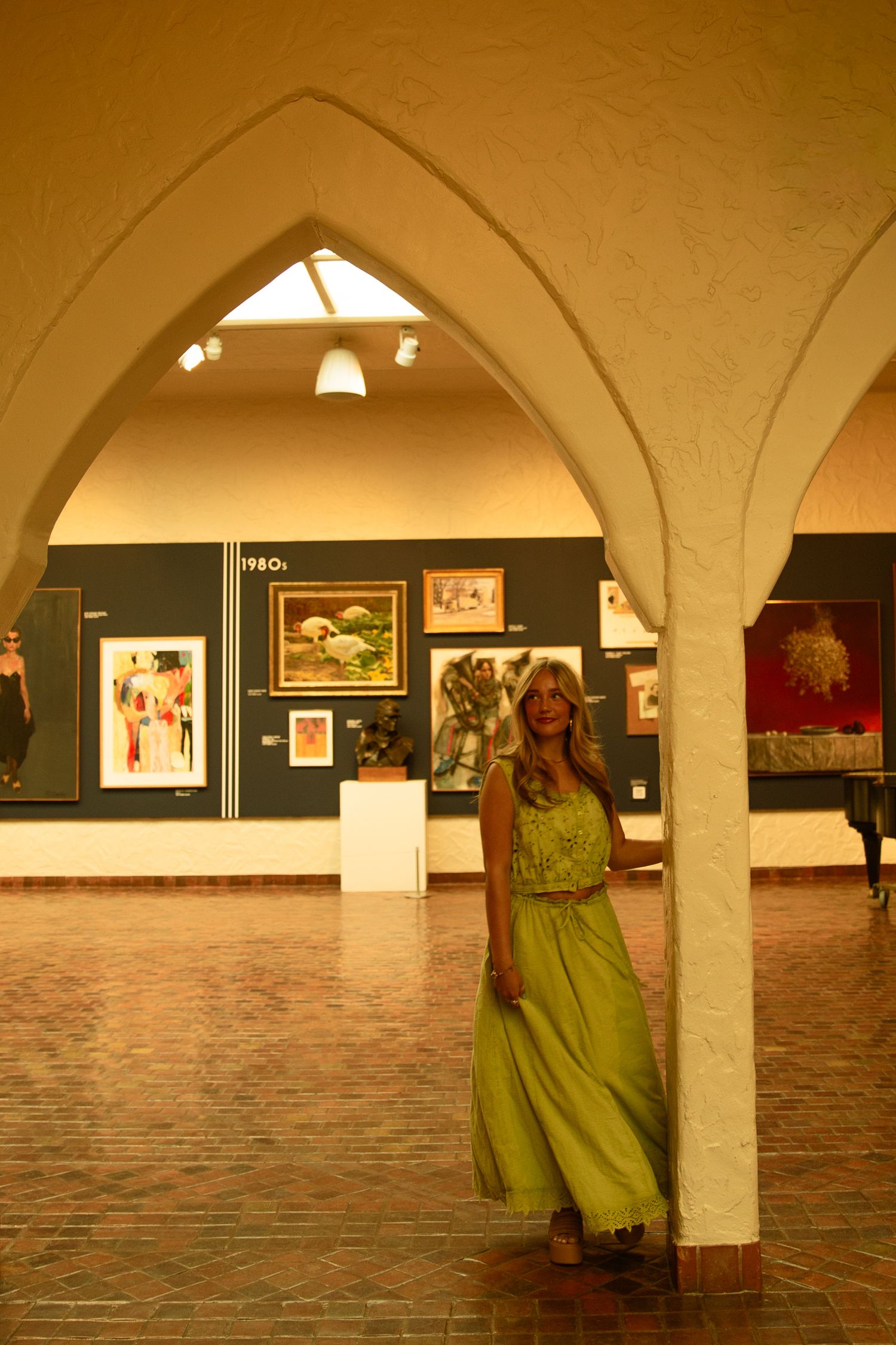 Woman in green gown poses by white arch column in art gallery with colorful paintings on display.