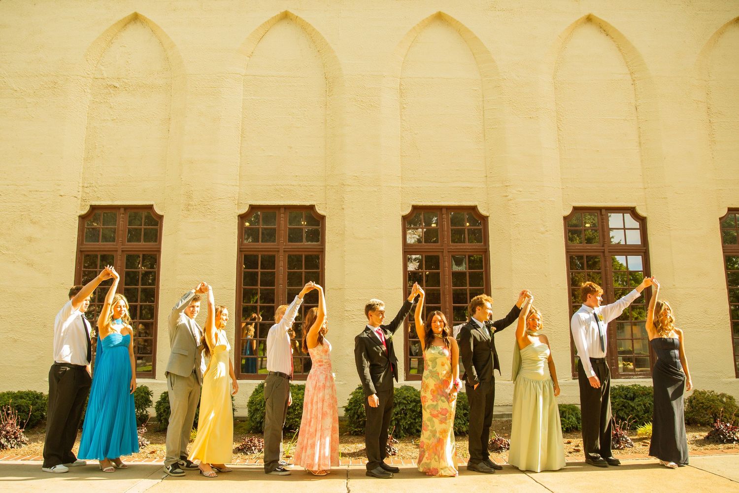 Wedding party in formal attire holding hands raised outside a yellow stone church building on a sunny day.