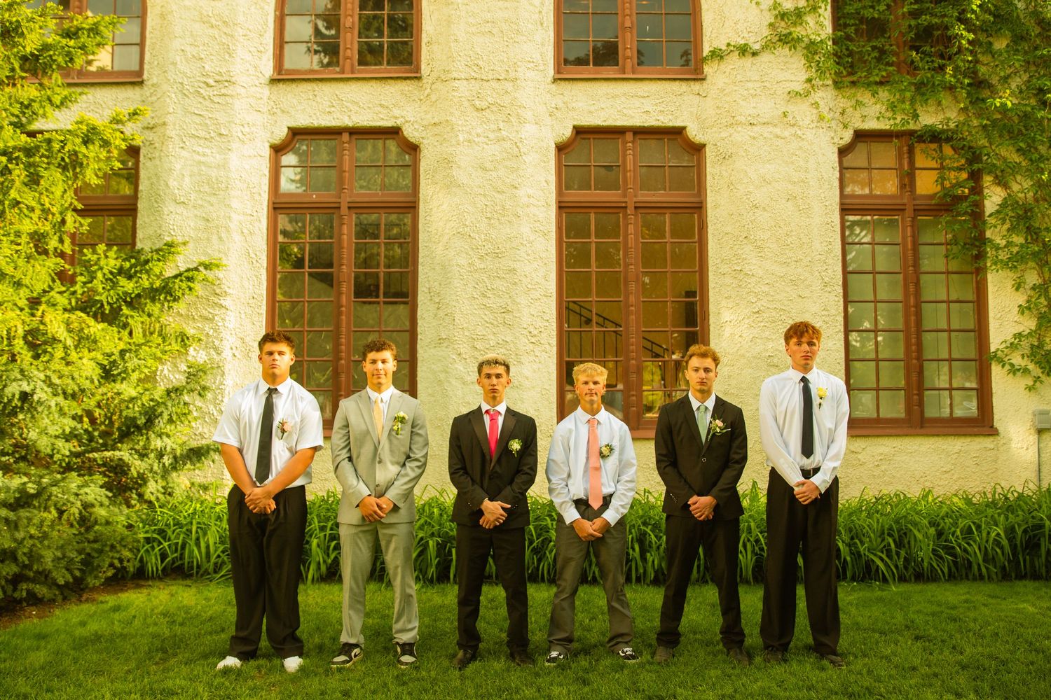 Six young men in formal suits and ties posing outdoors in front of a brick building with large windows.