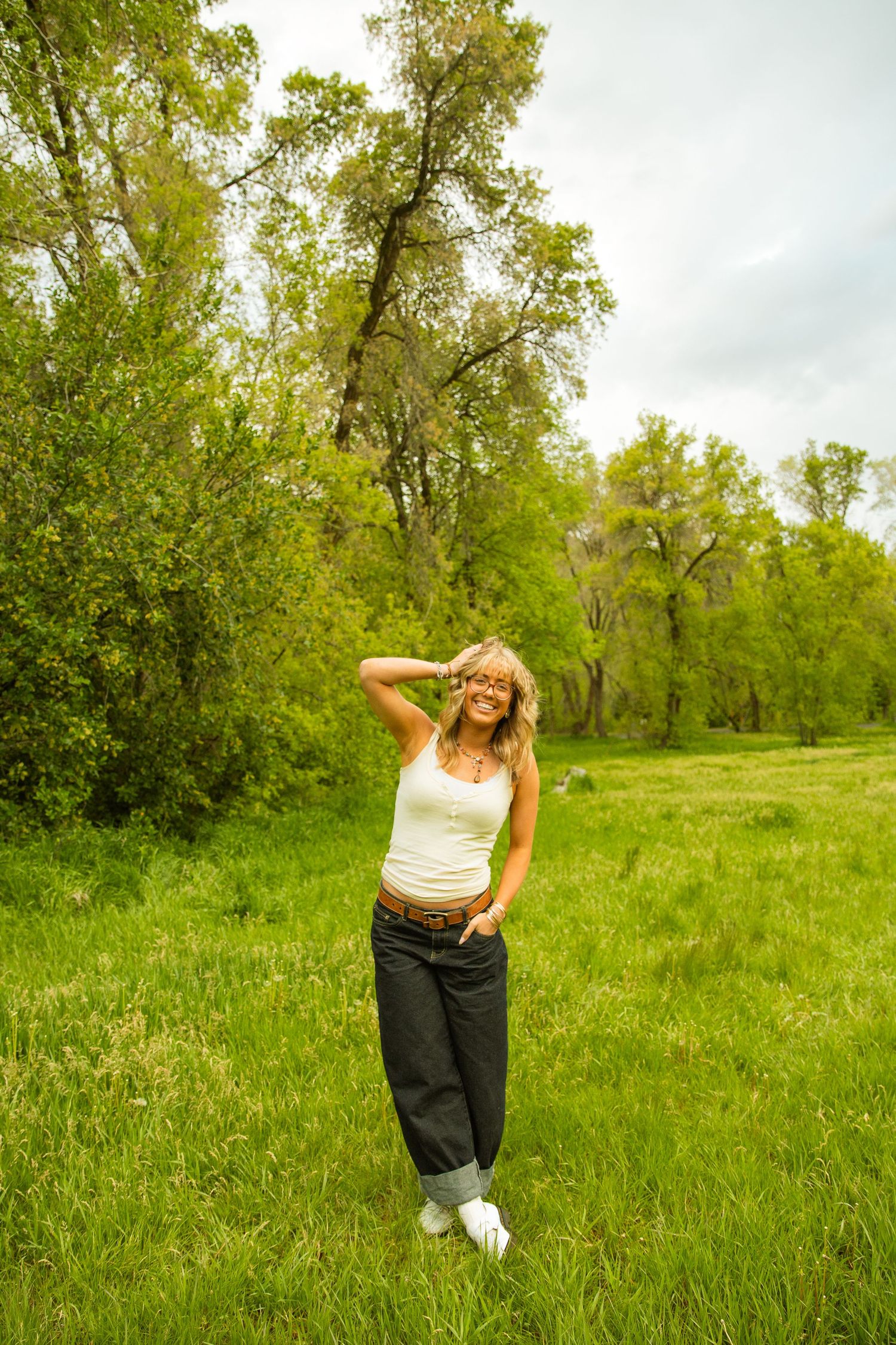 Smiling blonde woman in white tank top and dark jeans standing in lush green meadow with trees in background.