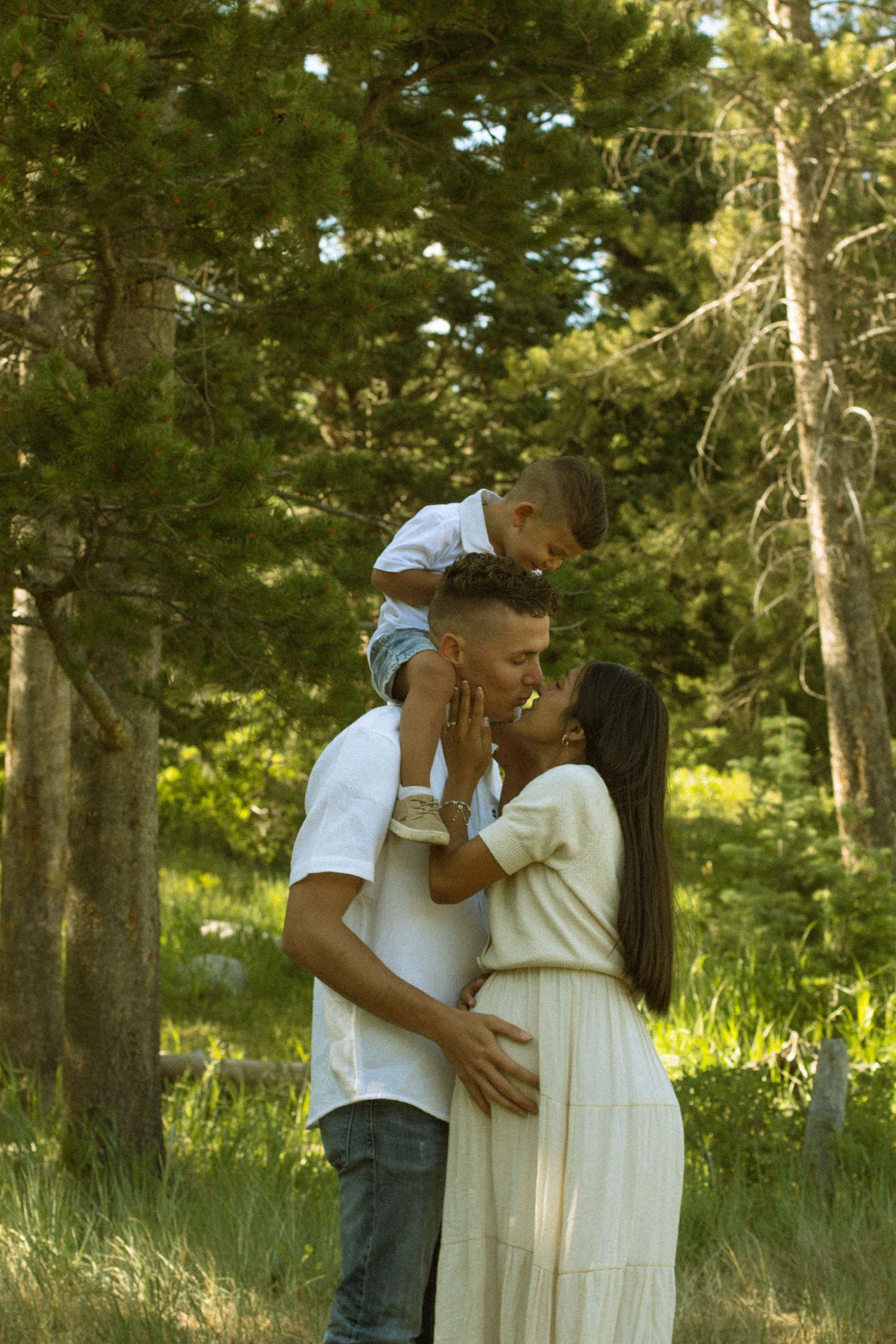 A romantic couple shares tender moments in a sunlit forest setting wearing white and blue casual attire.