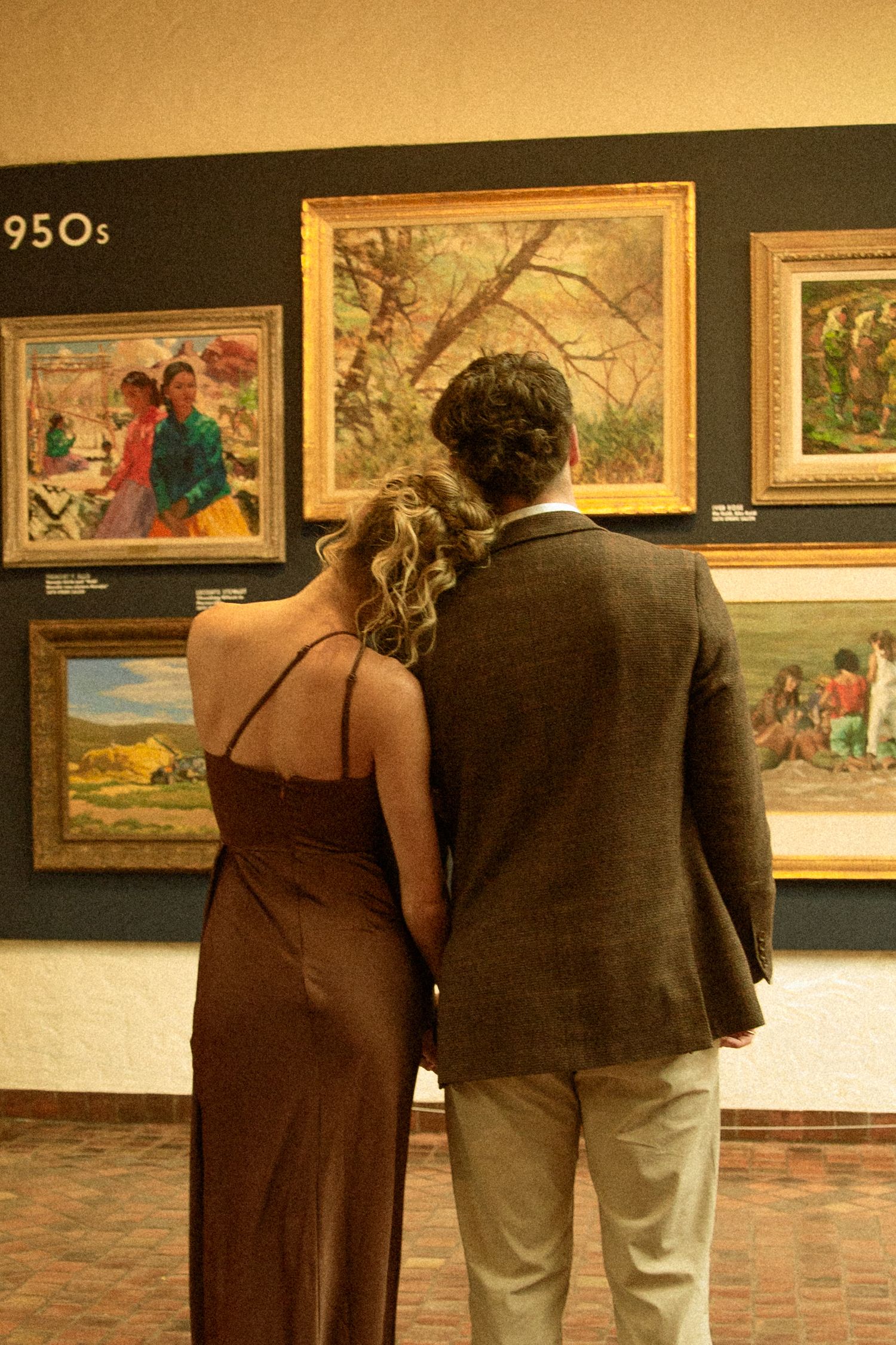 Two people viewing artwork displayed on gallery walls with warm lighting and brick flooring.