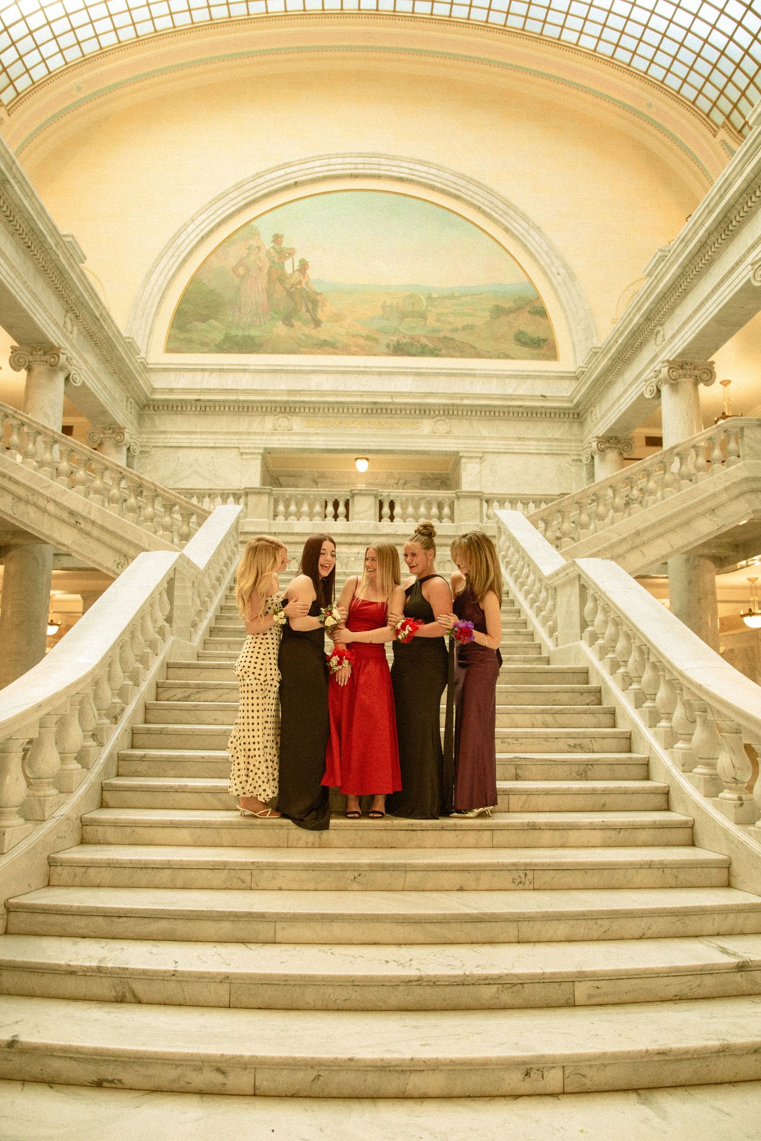Group in formal attire poses on grand marble staircase in elegant historic building with arched ceiling and ornate details.
