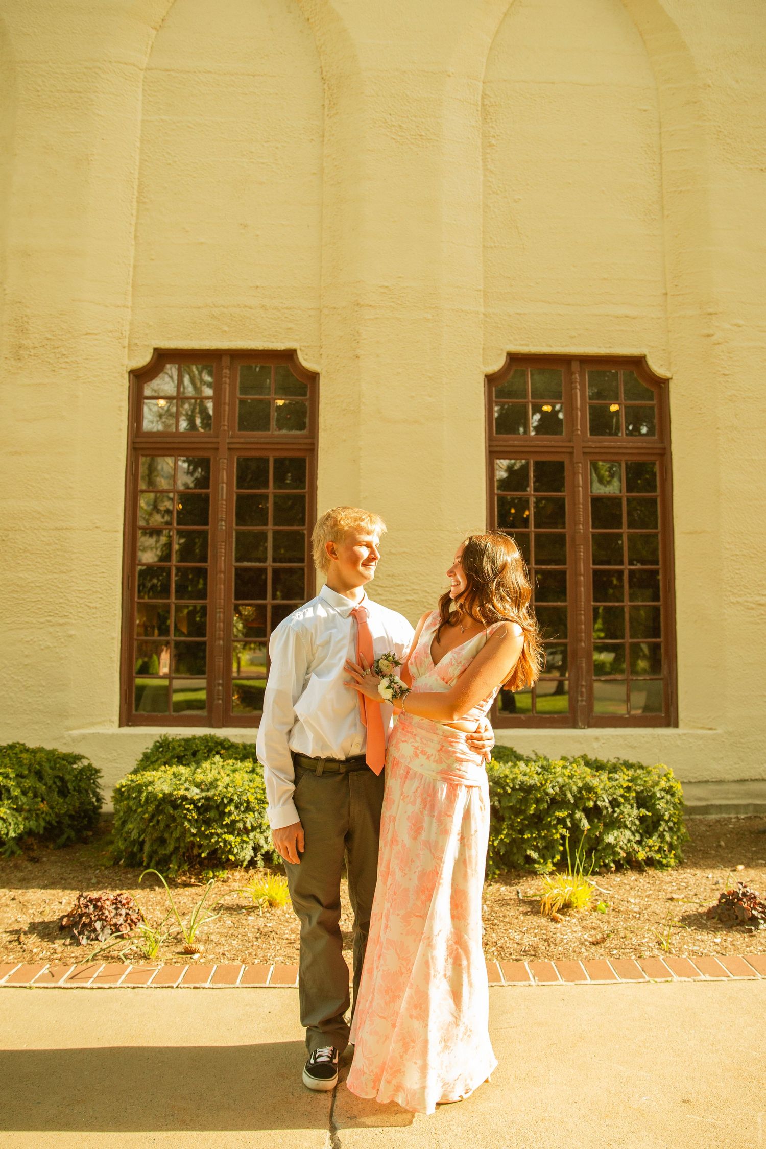 A couple in prom attire poses outside a yellow brick building, the young woman adjusting her date's coral tie.