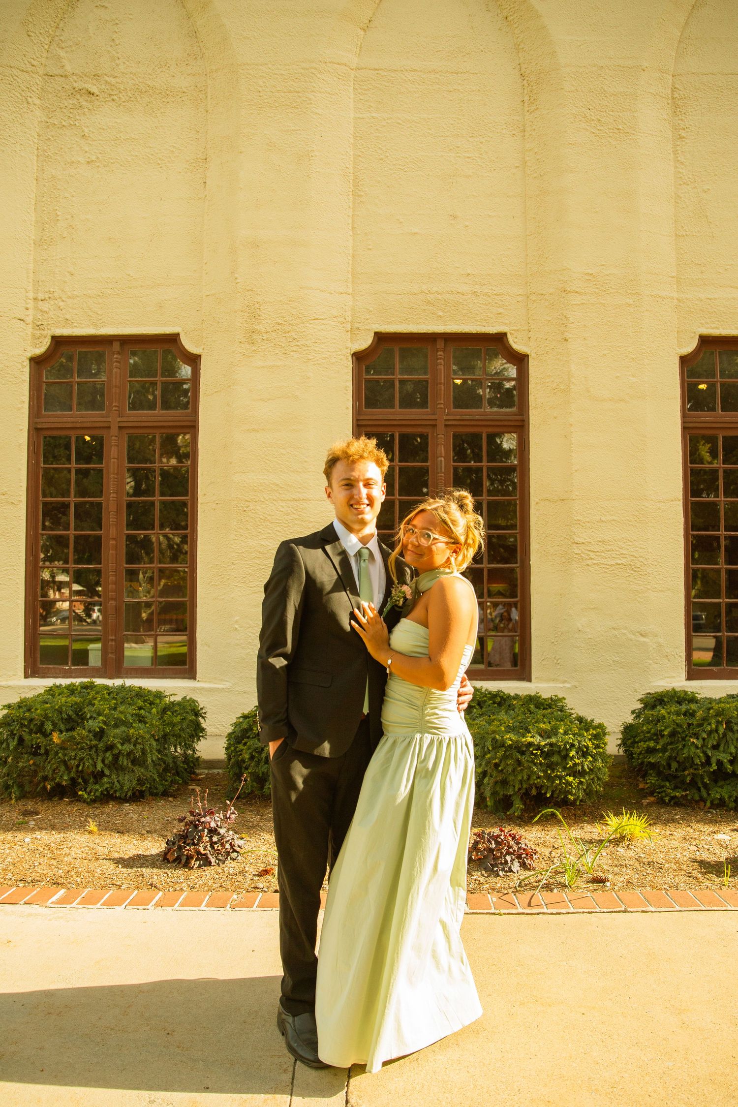A young couple dressed in formal prom attire poses outside a elegant building with arched windows.