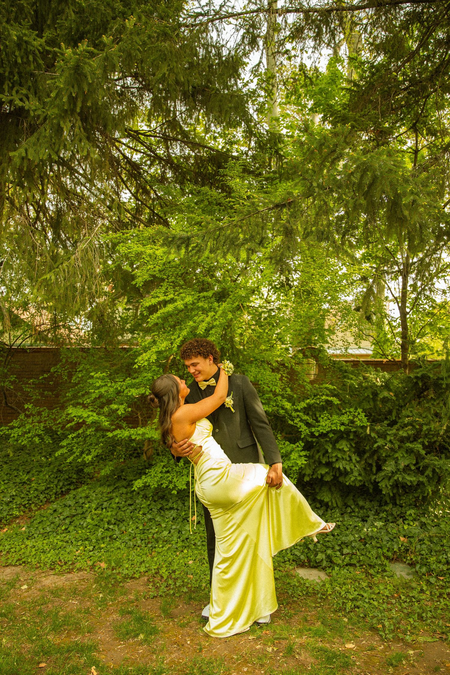 A couple shares a romantic dip kiss in a lush green garden, dressed in formal prom attire.