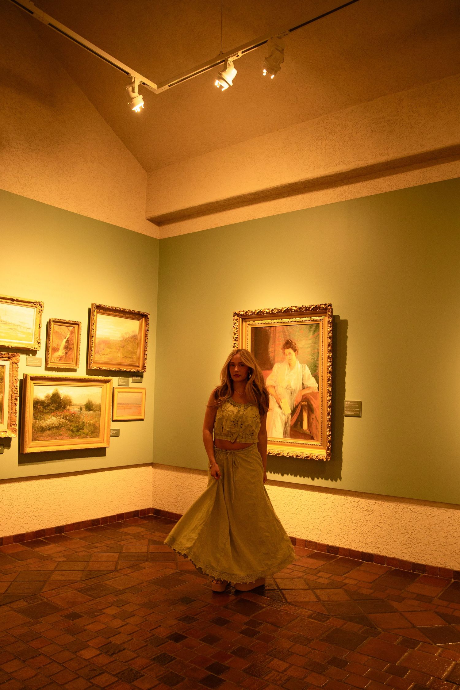 Woman in yellow gown posing in art gallery with framed paintings on green walls under warm lighting.