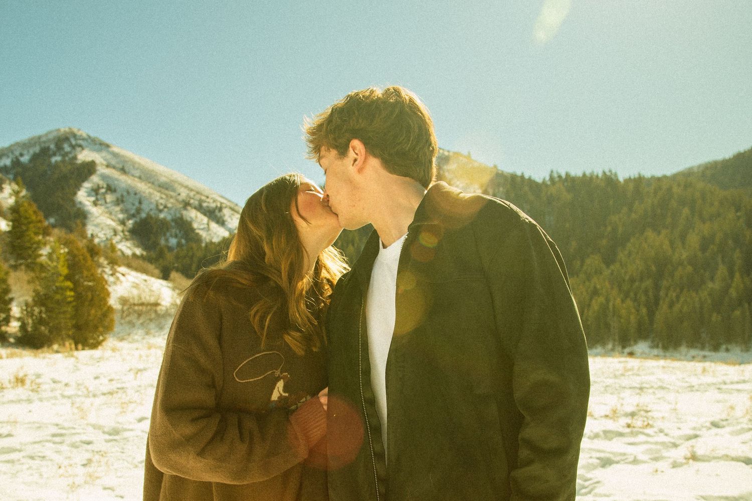 Two people embrace in a romantic winter scene with snow-covered mountains and sunlight creating a dreamy atmosphere.