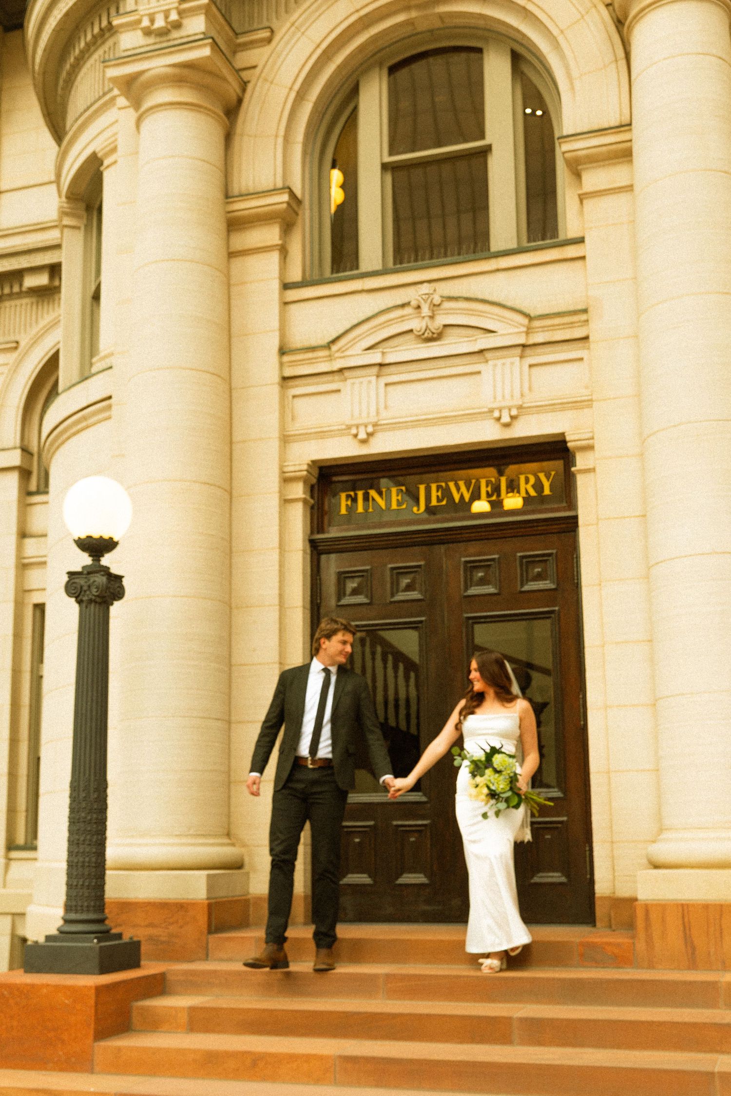 Wedding couple walking along a grand historic building facade with ornate columns and jewelry store signage.