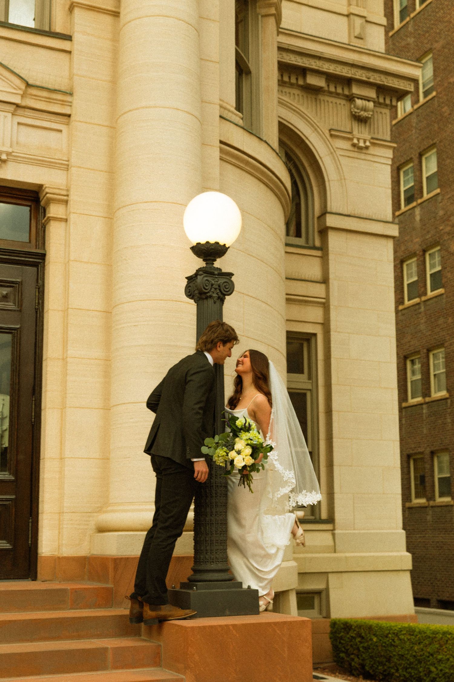 A couple shares a romantic moment on the steps of a historic building with ornate columns and a vintage lamp post.