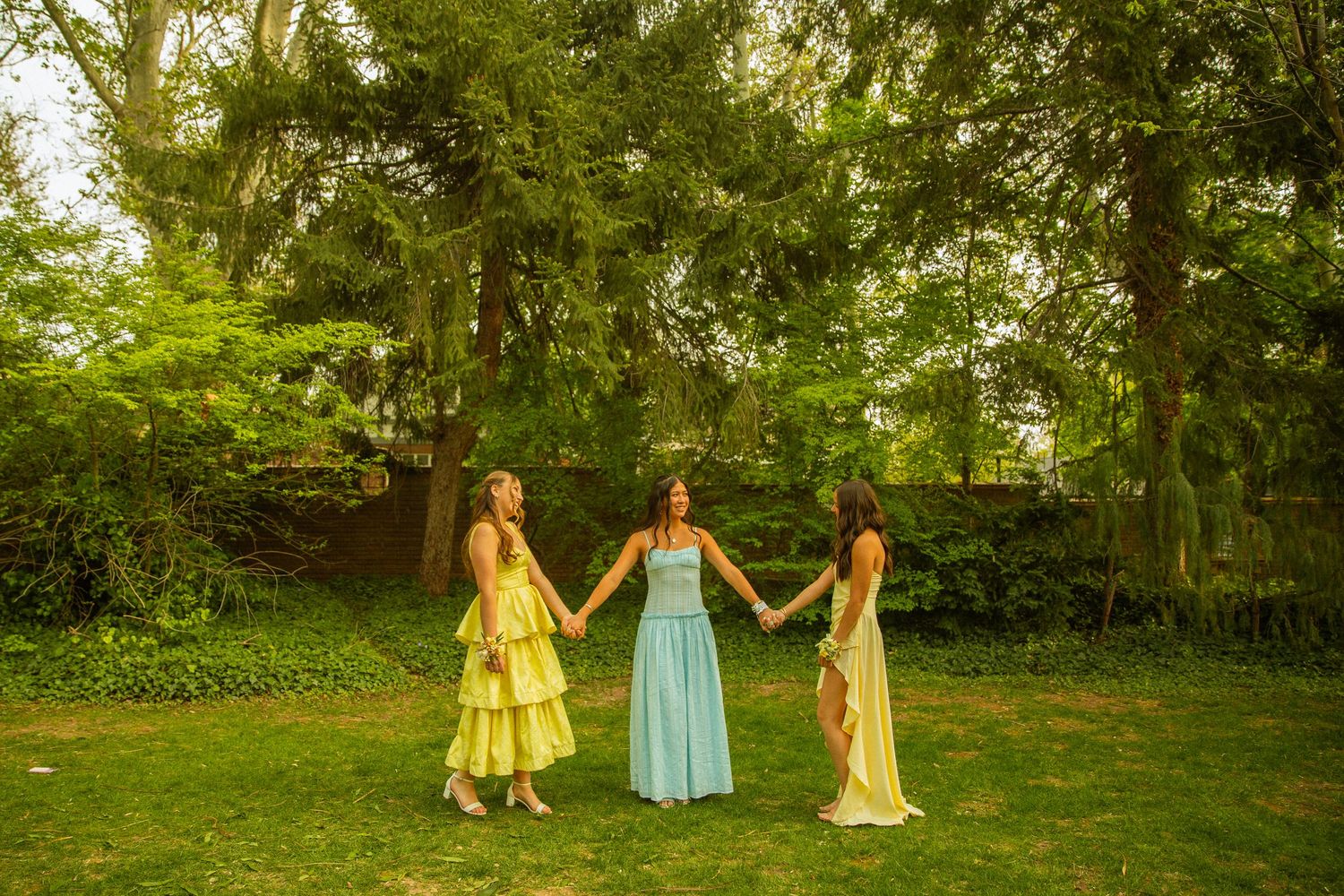 Three women in elegant pastel gowns hold hands in a lush green garden surrounded by tall trees.