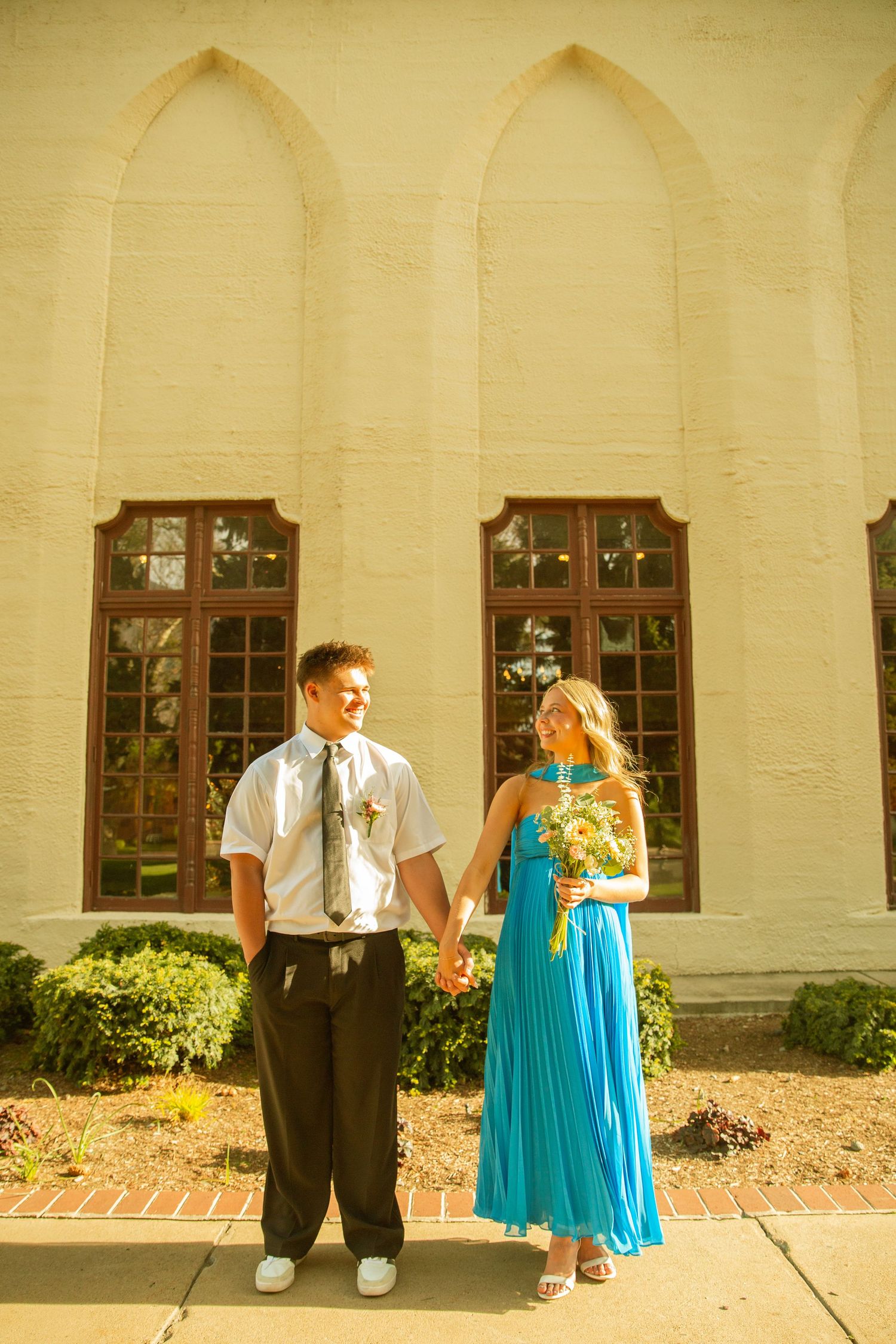 Couple holding hands in formal attire outside a church, woman in turquoise gown holding bouquet, man in white shirt and tie.