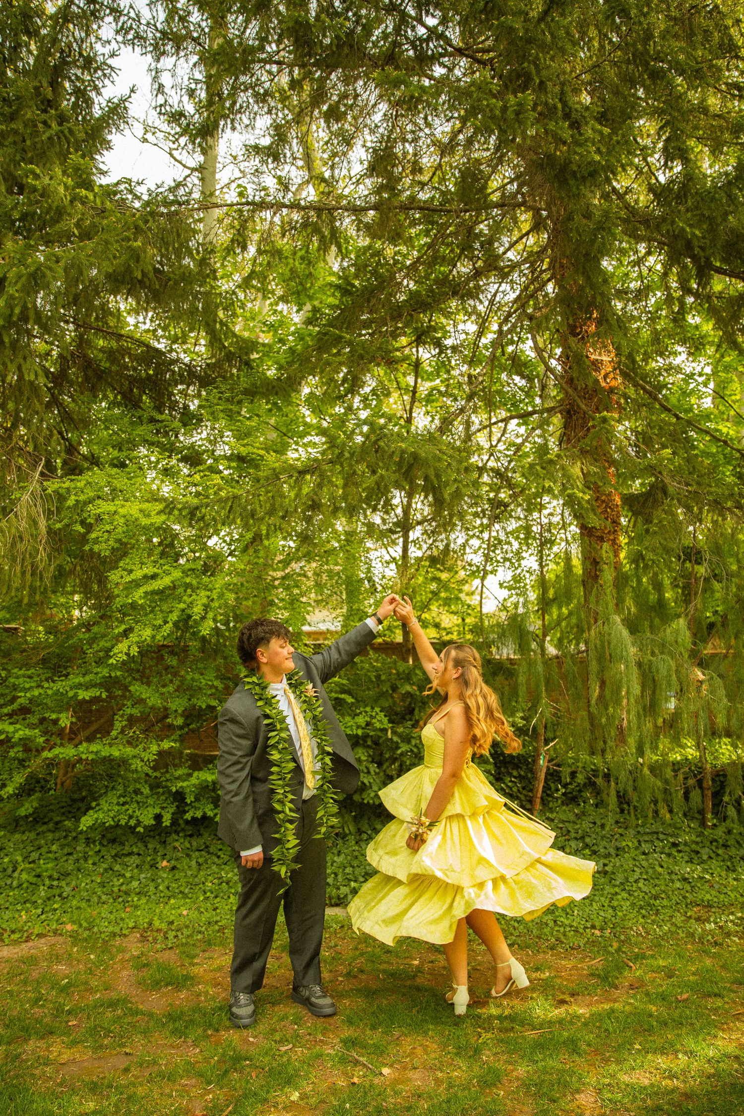 A couple dances outdoors, man in suit, woman in flowing yellow dress, surrounded by lush green trees.