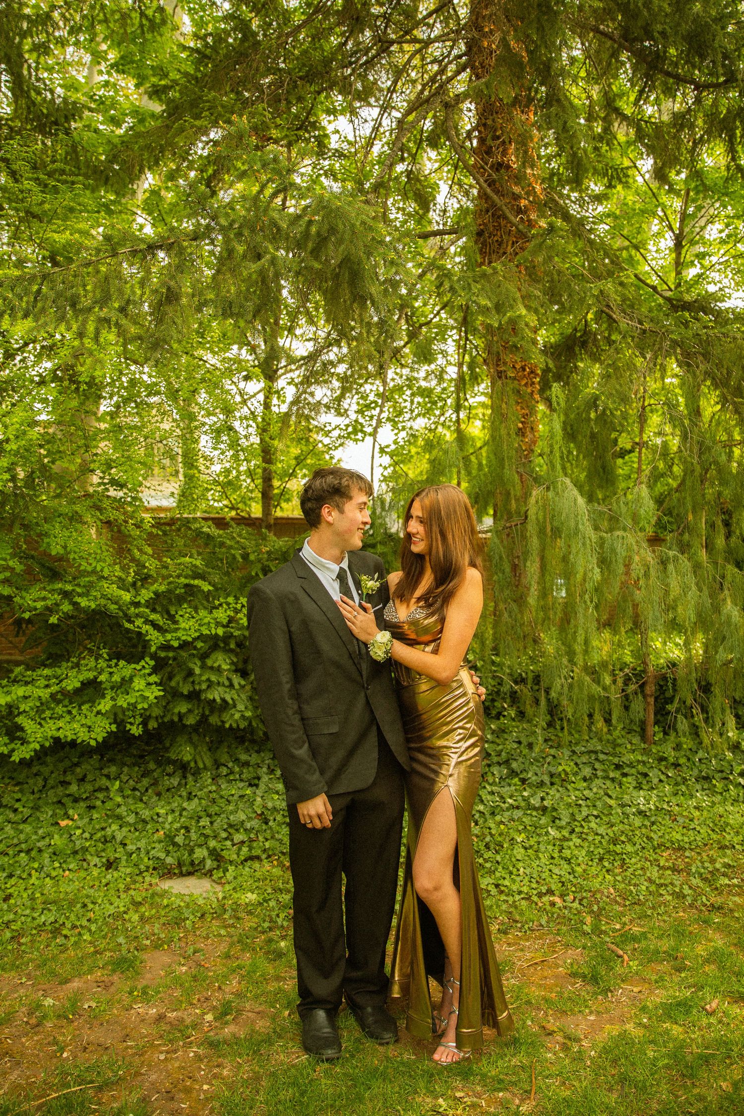 A couple in formal attire poses in a lush green garden, the woman in a gold dress and man in a dark suit.