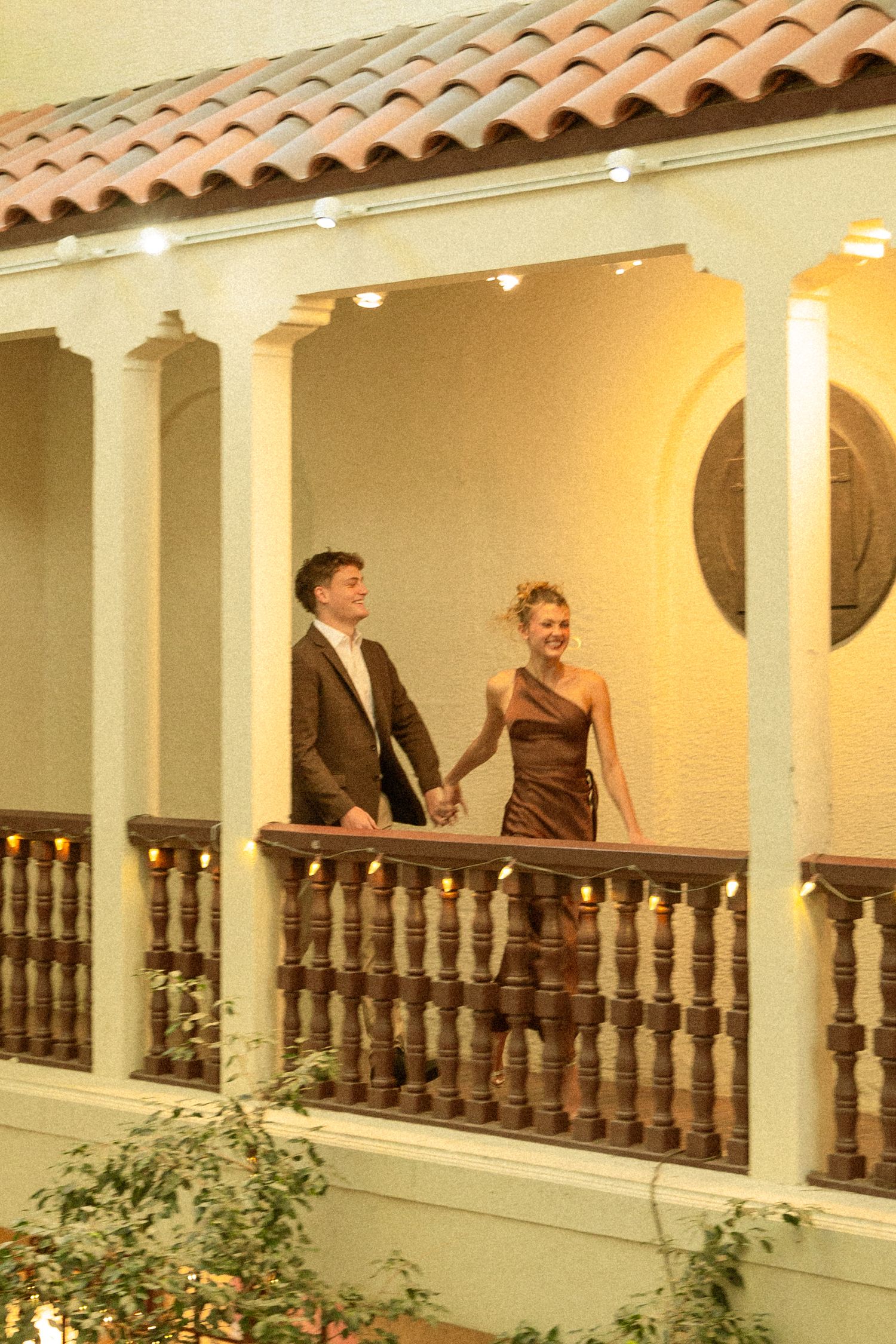 People gather on a Spanish colonial style balcony with white columns and terracotta roof tiles at sunset.