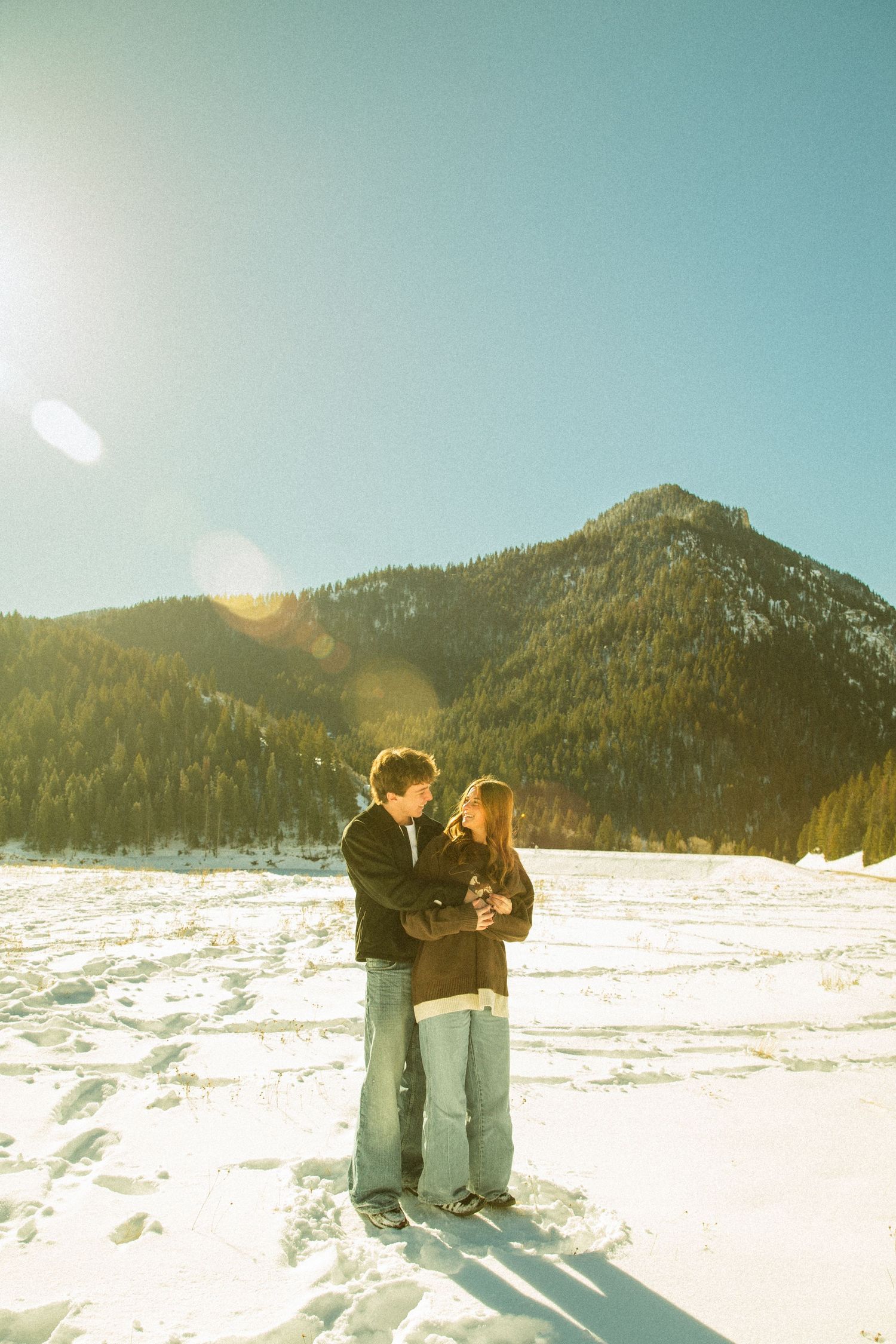 Two figures stand together in a snowy mountain landscape with sunlit peaks in the background.