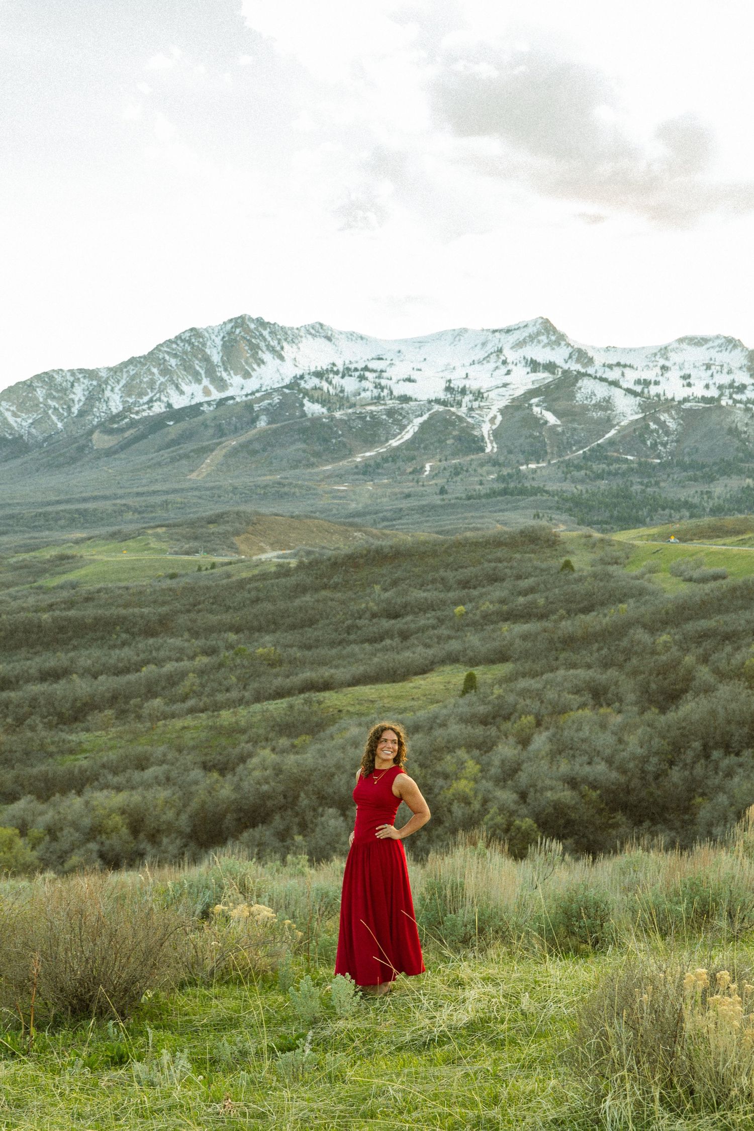 Woman in red dress poses in green mountain meadow with snow-capped peaks and cloudy sky backdrop.