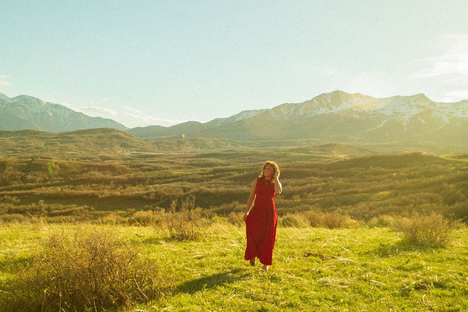 Woman in red dress standing in sunlit green meadow with mountain range backdrop on a clear day.