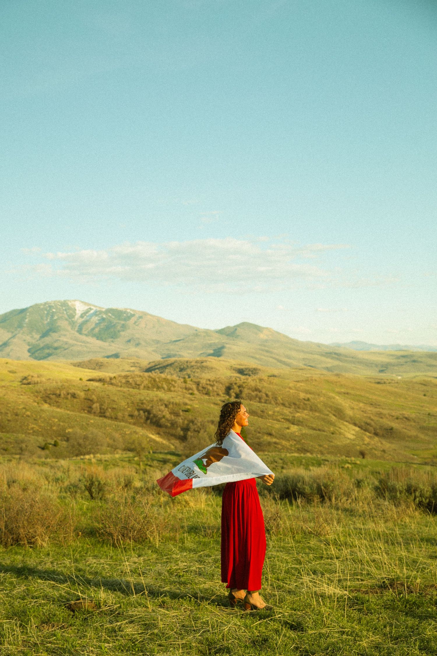 Woman in red skirt and white top holds a flag in a sunlit green mountain meadow under a clear blue sky.