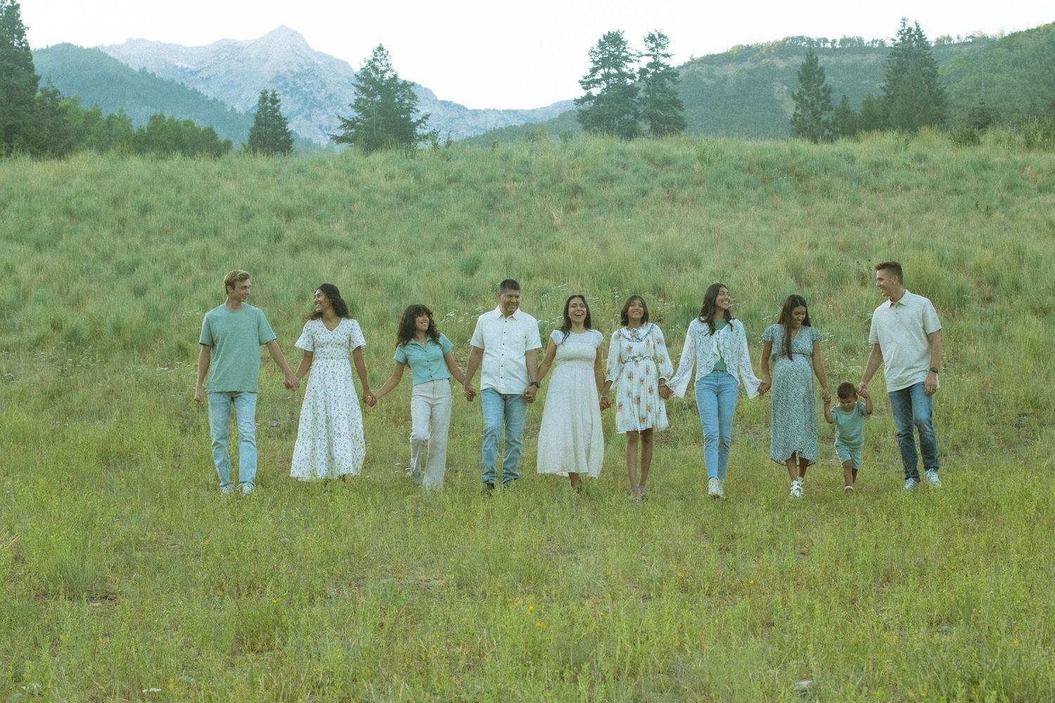 A group of people in white and blue clothing walk together through a green meadow with mountains in the background.