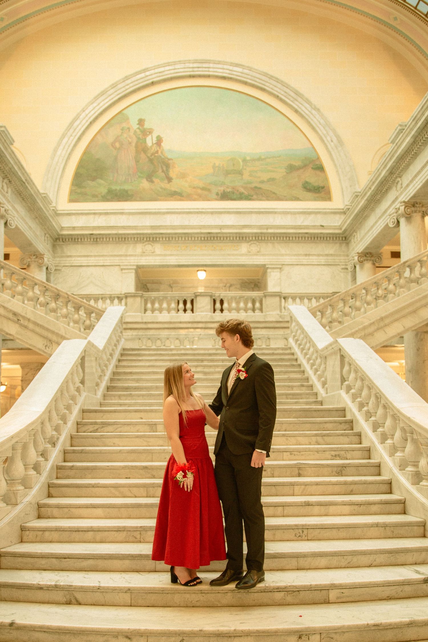 A couple in formal attire stands on an elegant marble staircase with ornate architecture and a painted mural above.