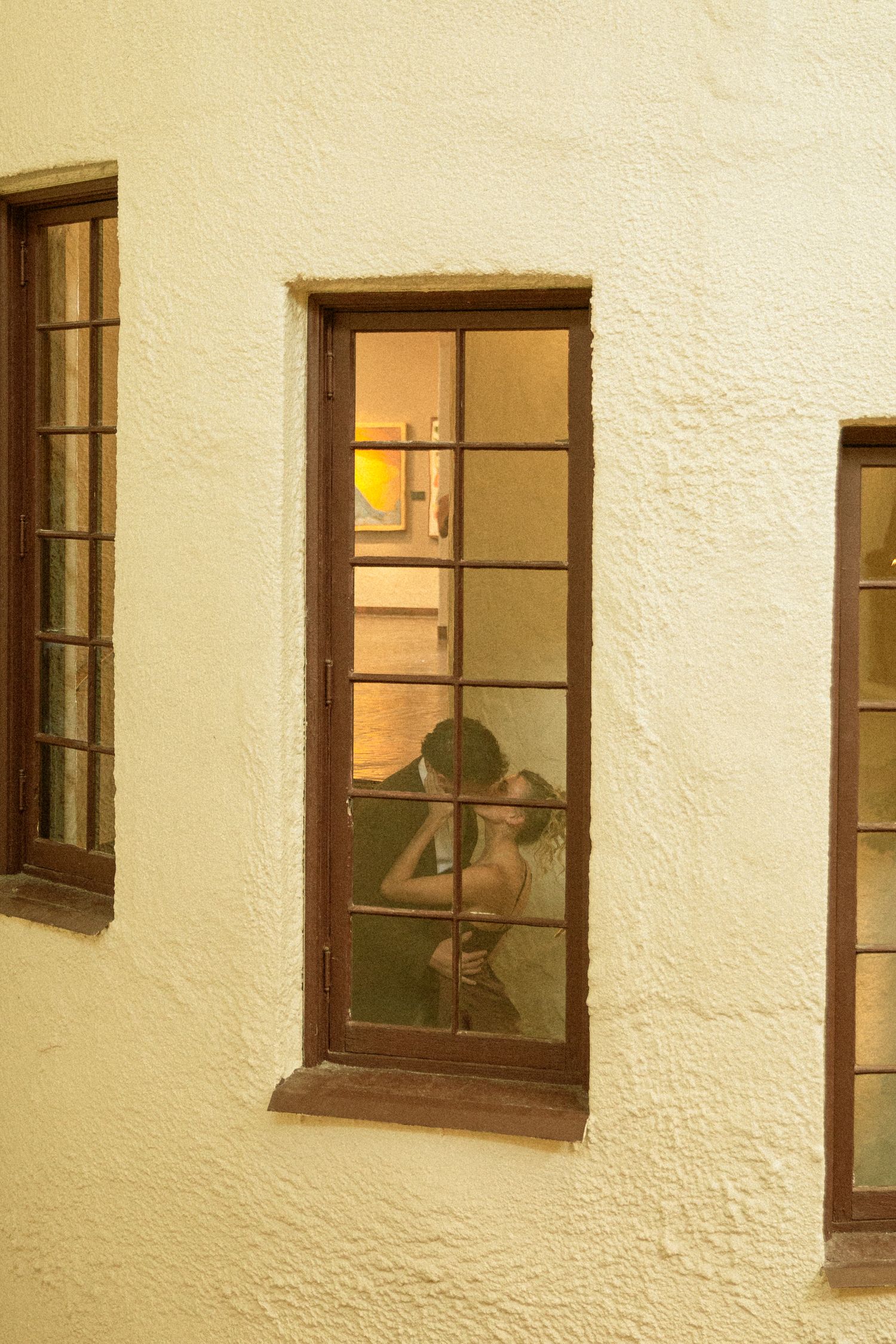 A row of illuminated windows on a cream-colored building exterior shows silhouettes moving inside at dusk.