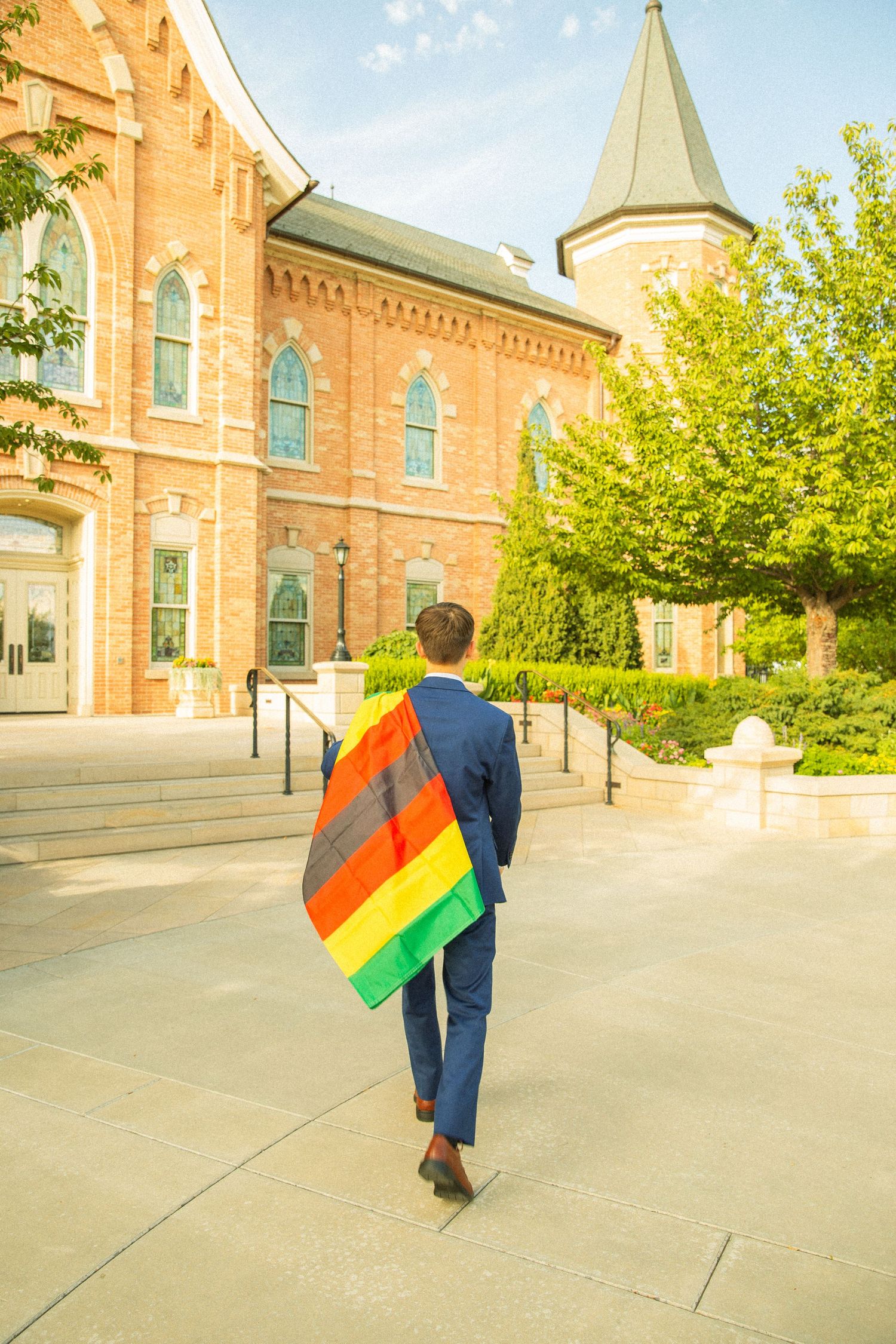 A person wearing blue clothing carries a pride flag while walking outside a historic brick church building with a spire.