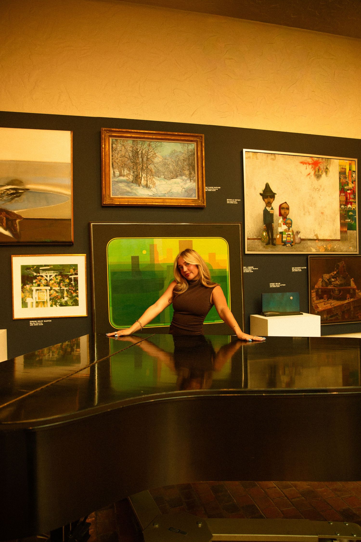 Blonde woman posing in front of colorful artwork display in a gallery, surrounded by paintings on a dark wall.