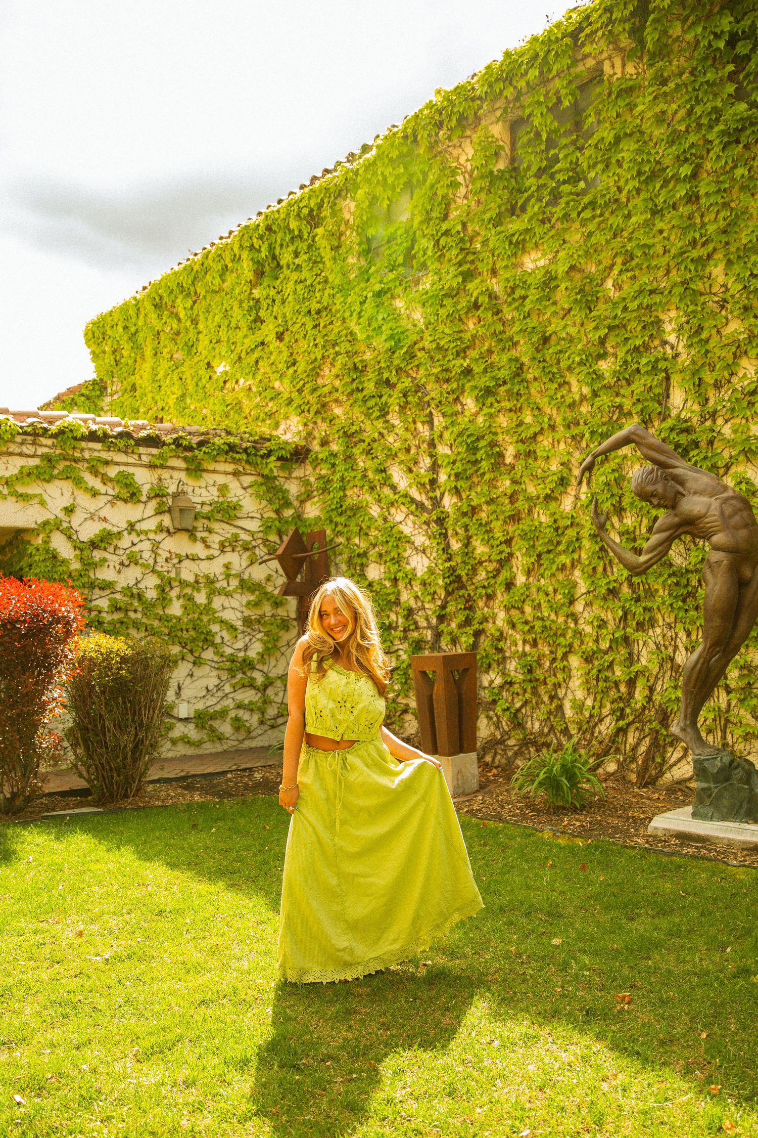 Woman in yellow maxi dress standing in lush garden with ivy-covered wall and bronze sculptures.