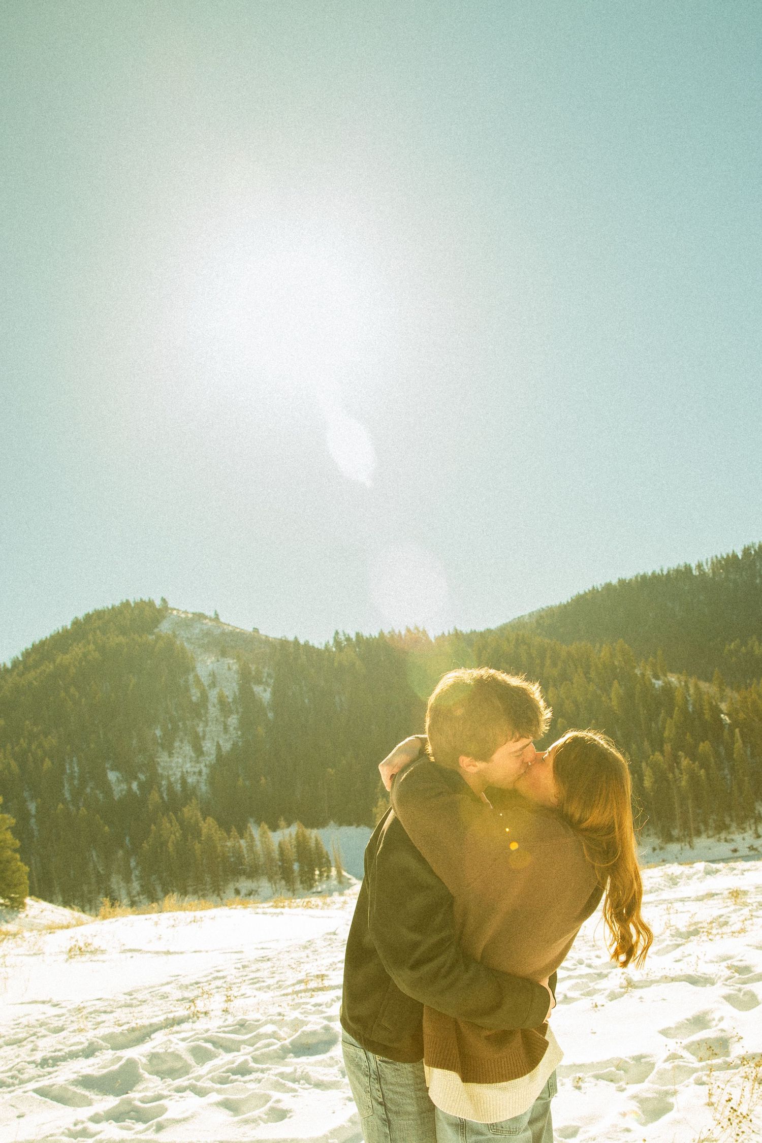 A couple shares a romantic kiss against a snowy mountain backdrop with sunlight creating a dreamy winter atmosphere.