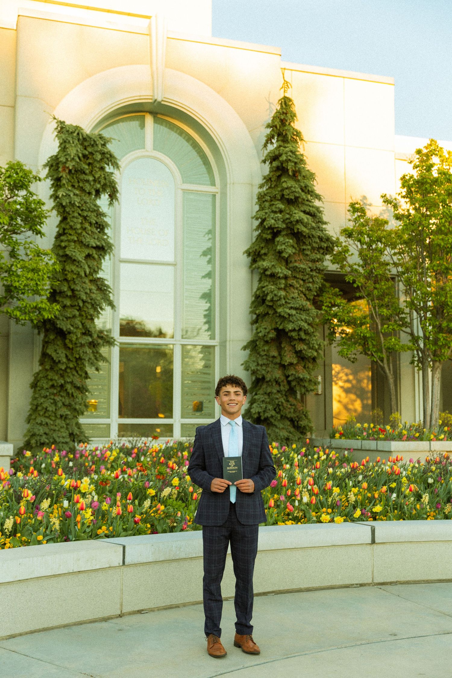 Young man in navy suit holding scriptures stands before a sunlit building with evergreen trees and colorful tulips.