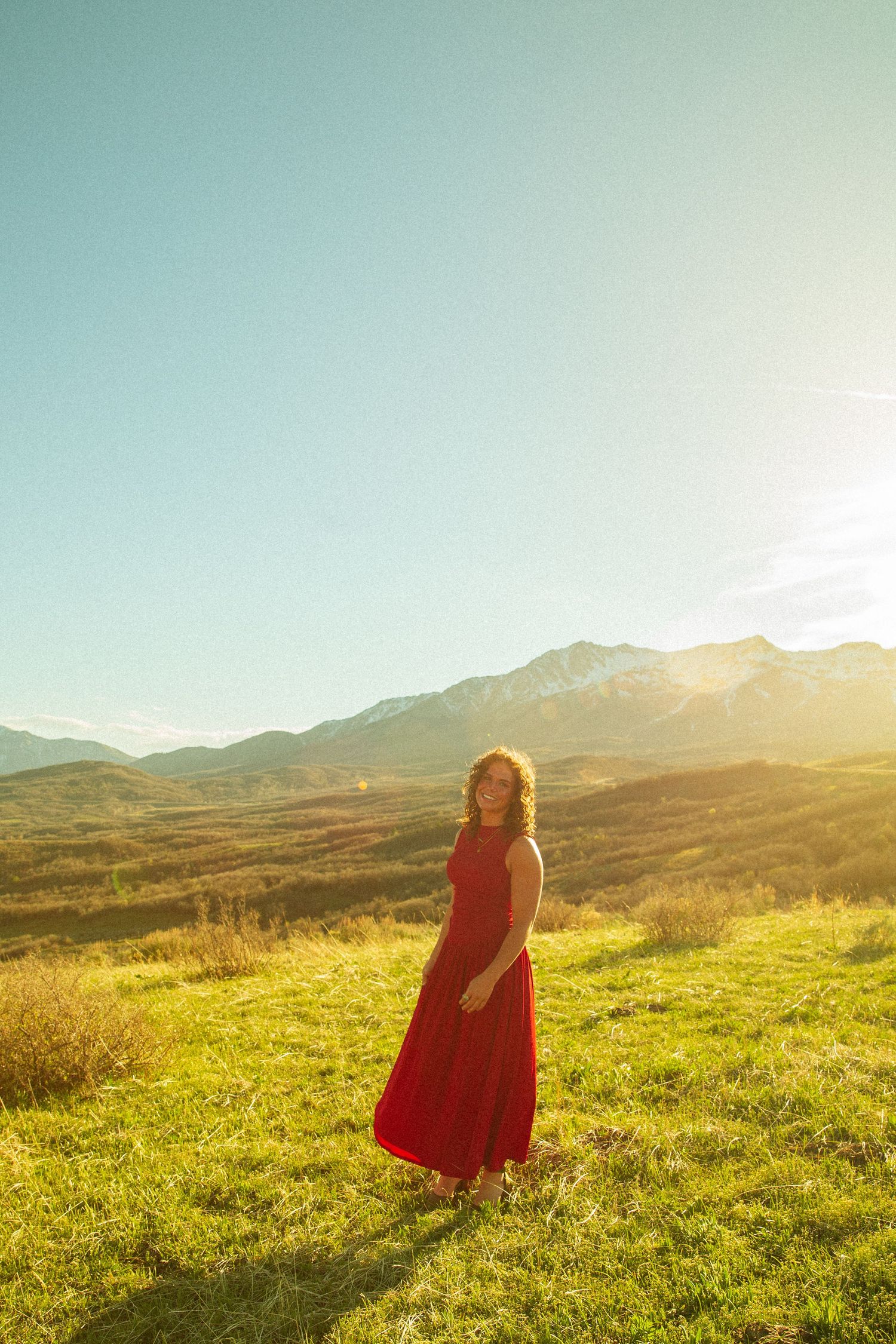 Woman in red maxi dress standing on green meadow with mountain landscape and golden sunset sky behind her.