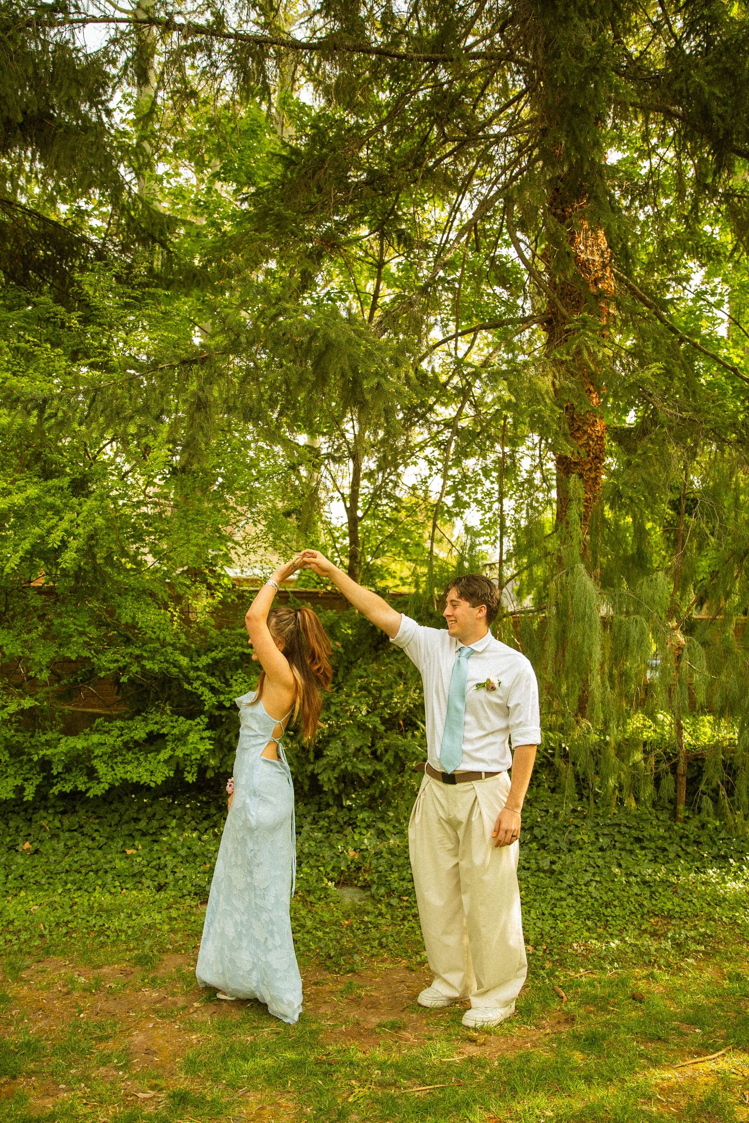 A bride and groom dancing outdoors under lush green trees, the groom spinning his bride in a white lace wedding dress.