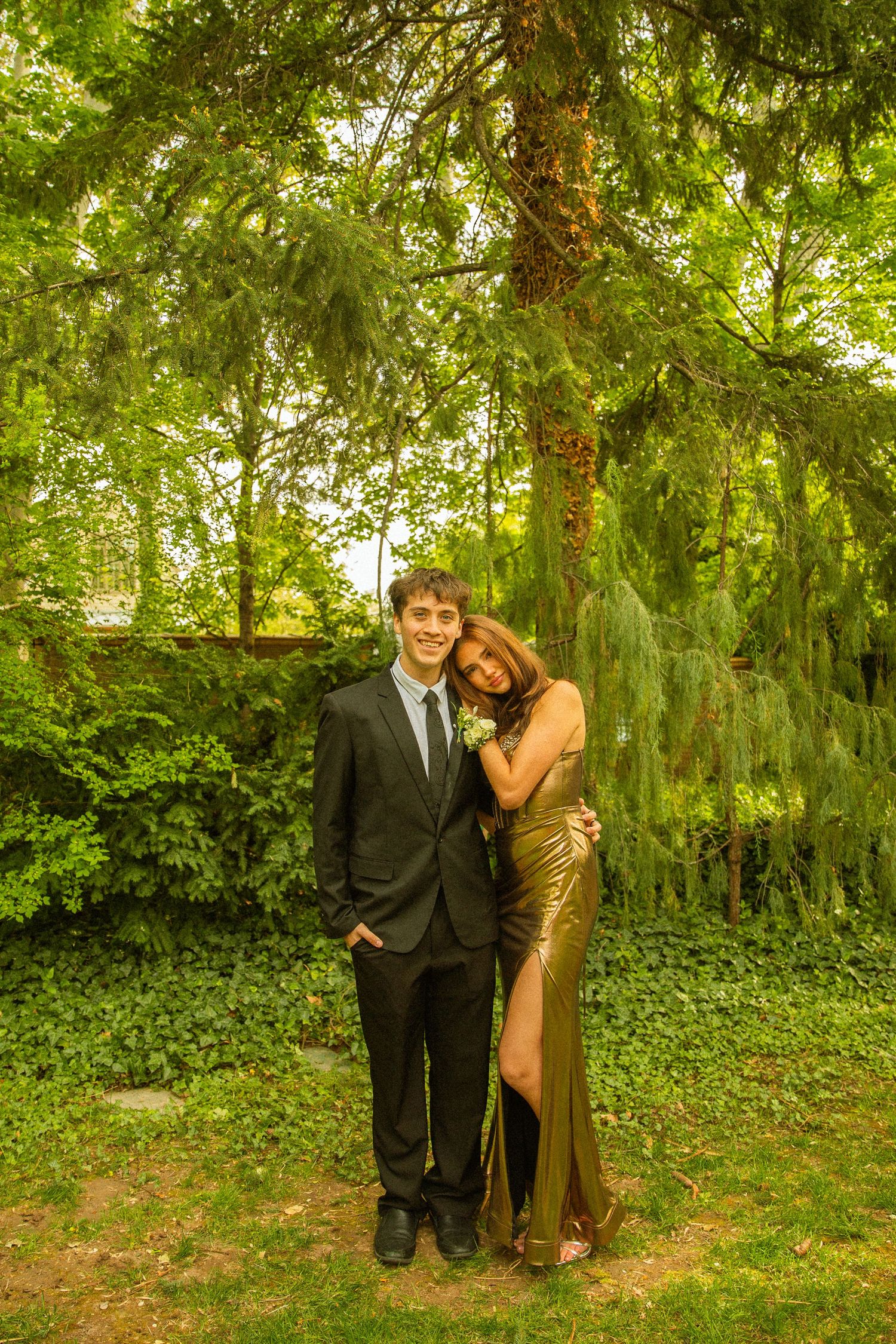 A young couple poses in a lush garden, wearing formal prom attire, a black suit and gold gown.