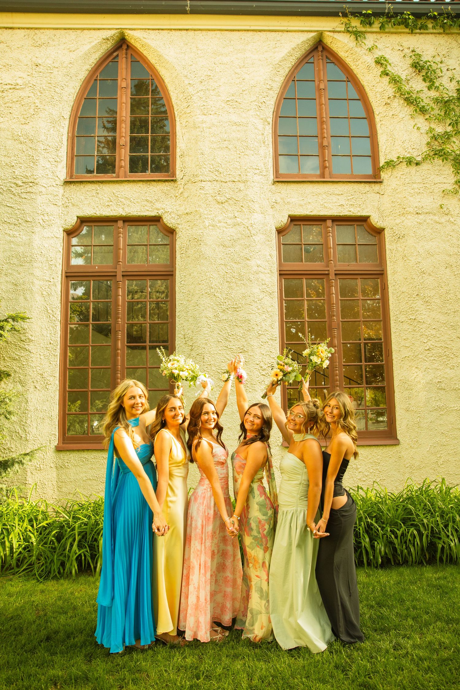 Six bridesmaids in colorful mismatched gowns raising bouquets outside a stone church on a sunny day.