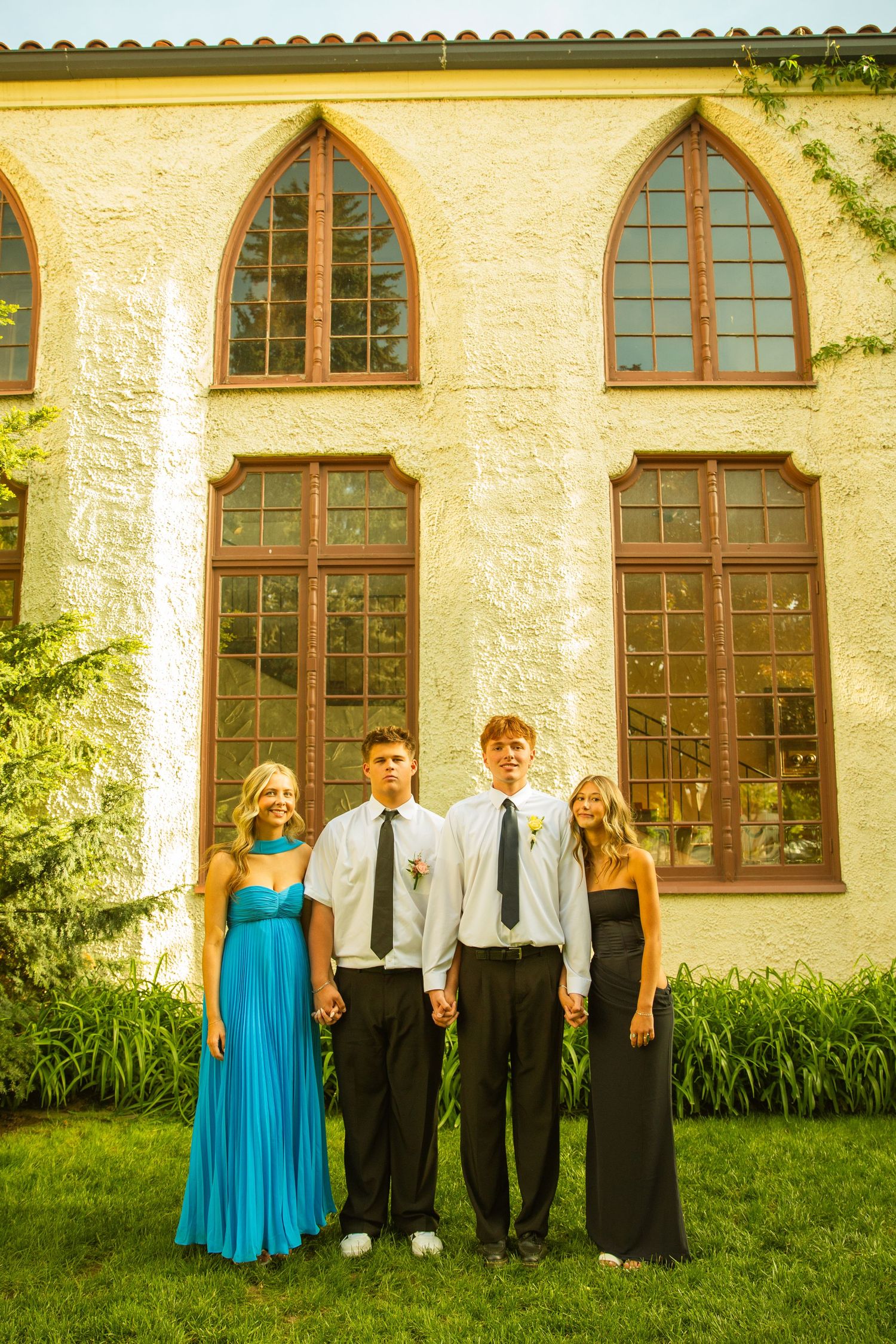 Two teen couples in formal attire pose in front of a stone building with gothic arched windows at prom.