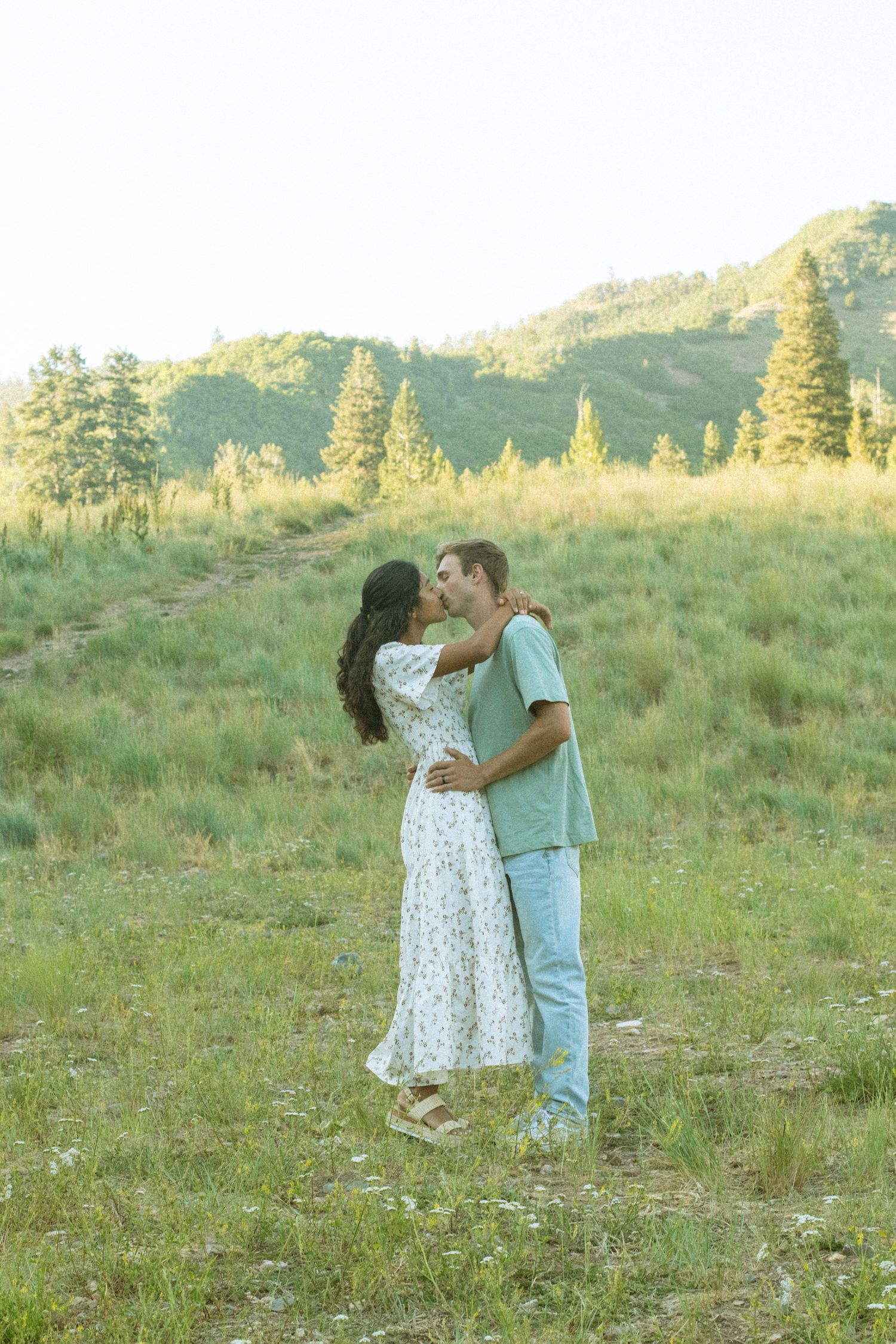 A couple shares a romantic moment in a sunlit meadow with wildflowers and mountains in the background.
