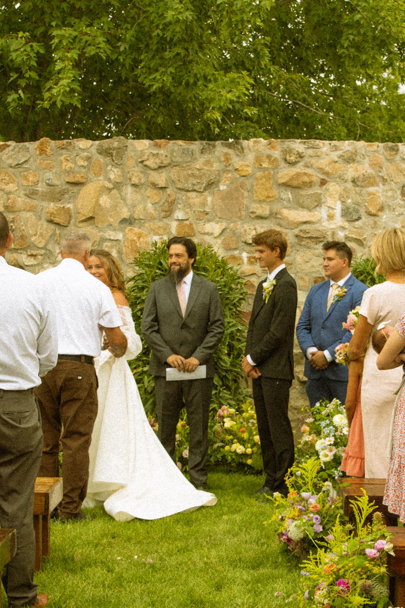Wedding ceremony guests lined up along stone wall with colorful garden flowers.