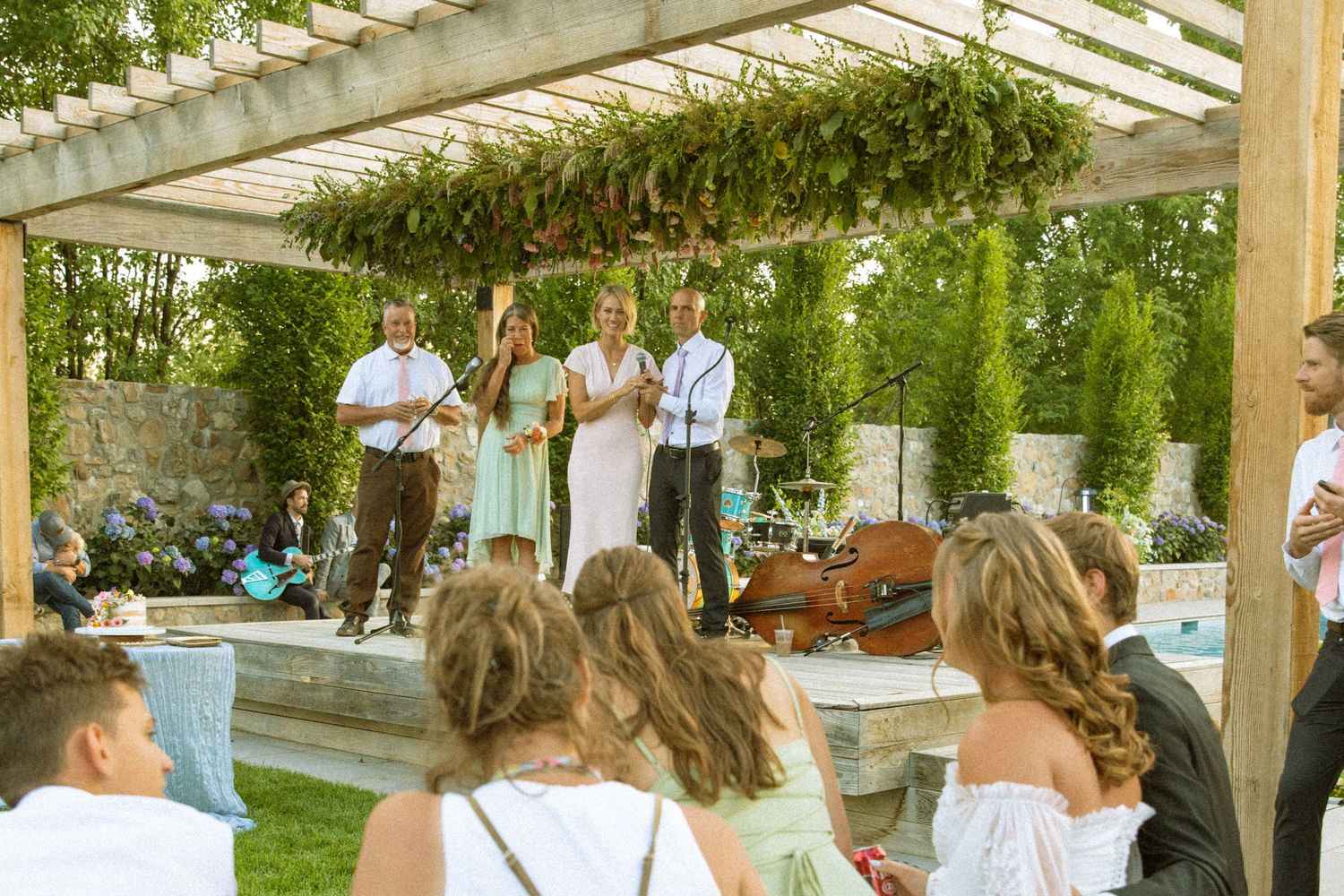Wedding guests mingle under a wooden pergola decorated with greenery.