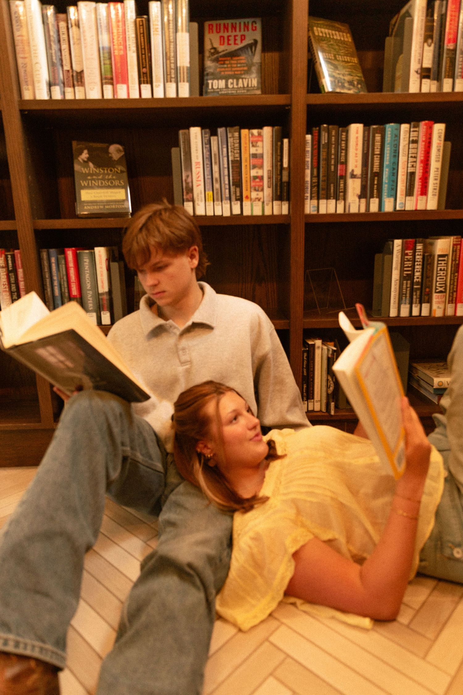 A person lies on the floor of a library reading a book while surrounded by wooden bookshelves filled with books.