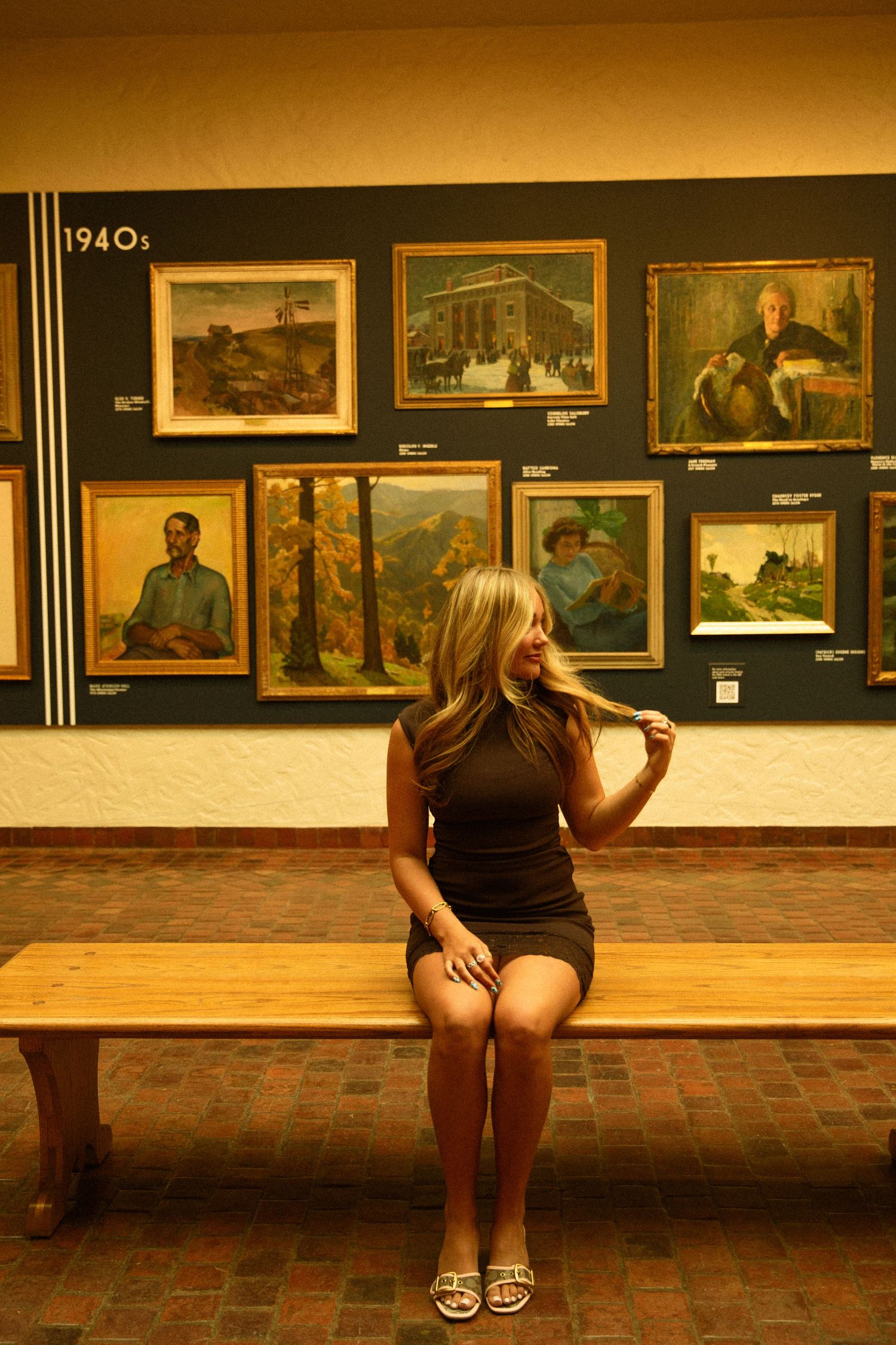 Blonde woman in black dress sitting on wooden bench in art museum gallery with 1940s paintings on blue wall.